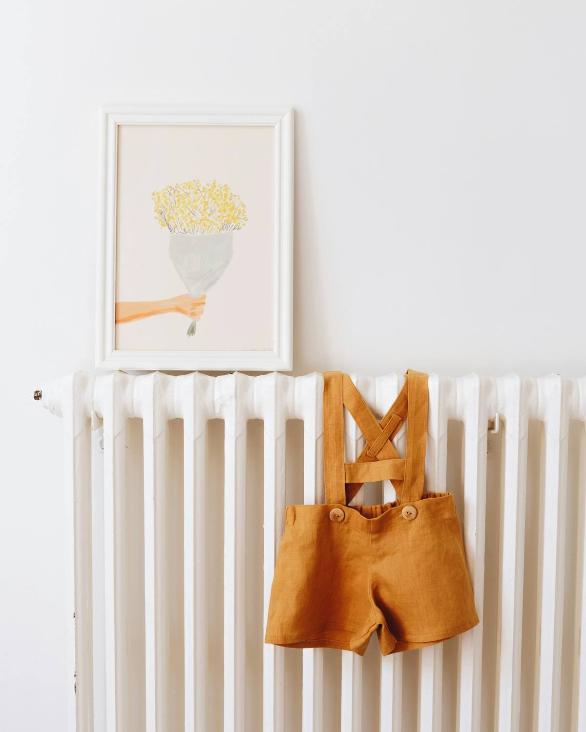 A child's mustard dungarees hanging on a radiator below a framed print