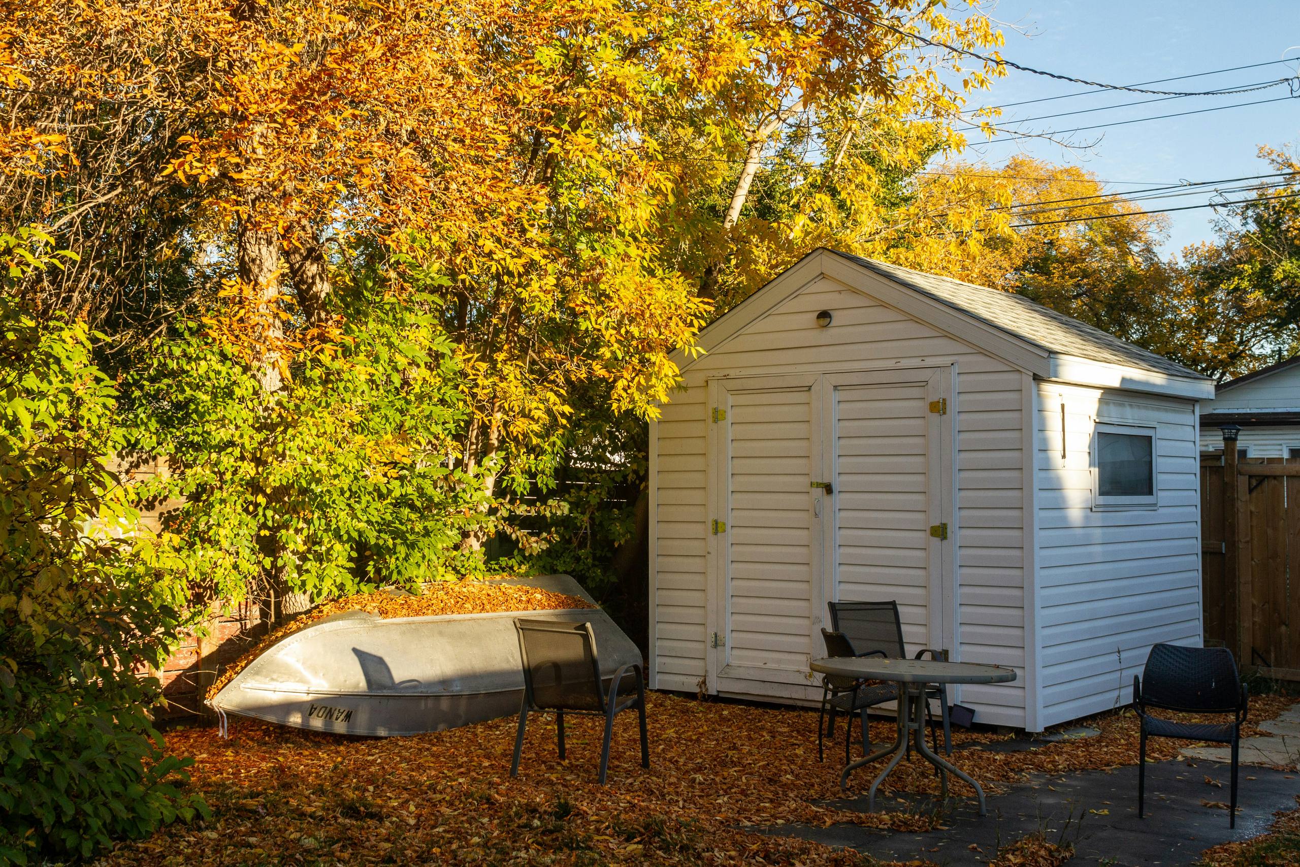 A white garden shed among autumn trees, with an upturned boat beside it