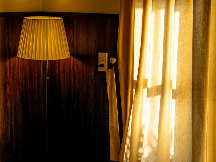 A bedroom at dusk lit by a single lamp, with sunlight through gold curtains
