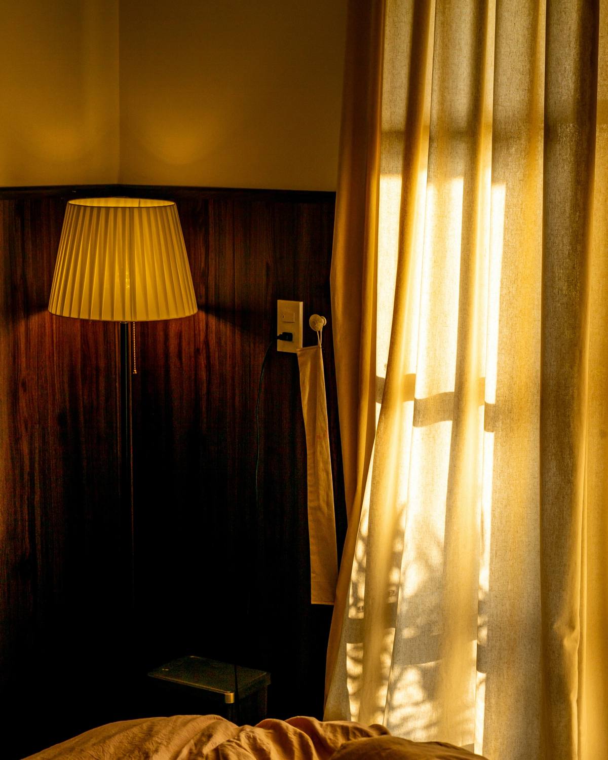 A bedroom at dusk lit by a single lamp, with sunlight through gold curtains