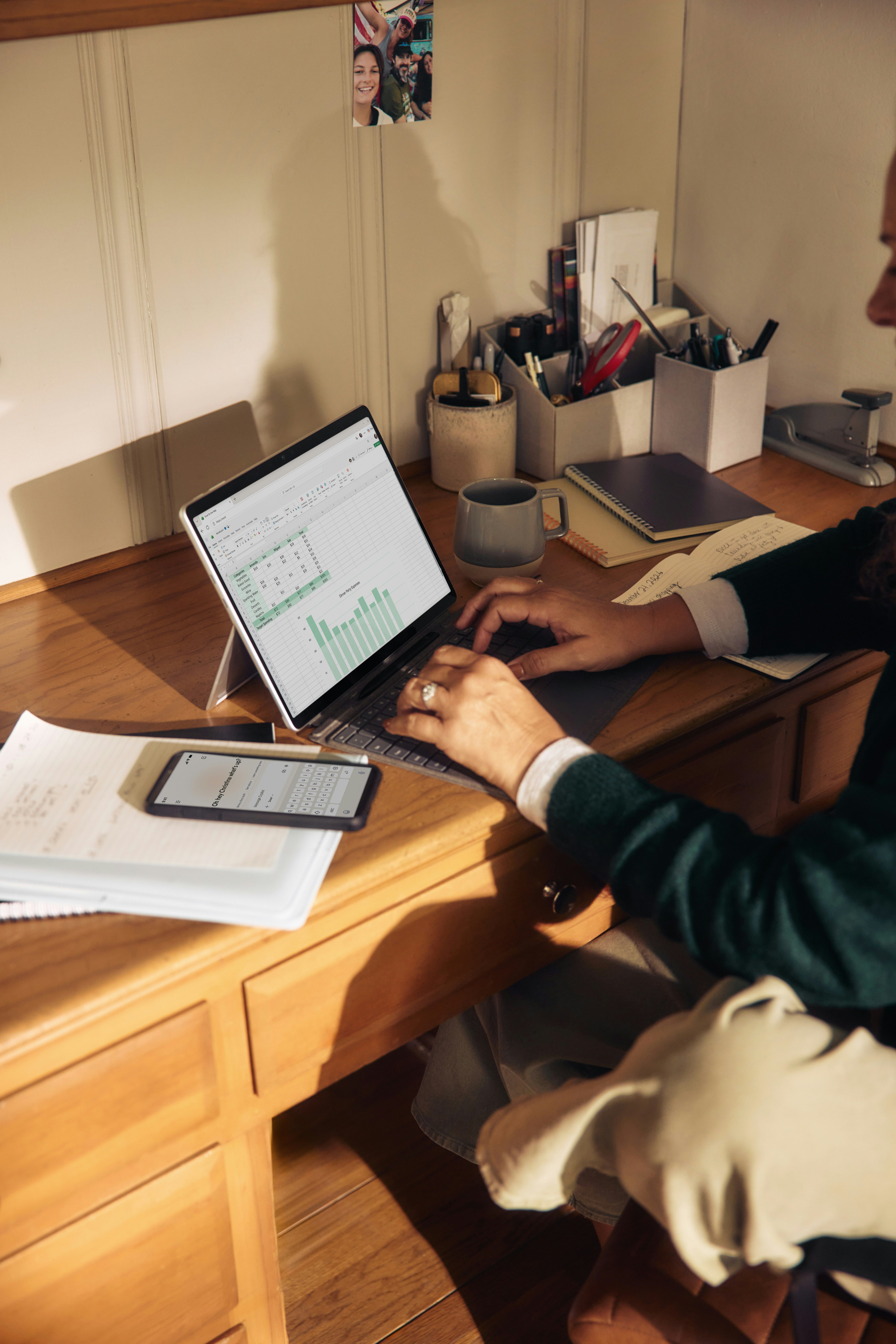 Someone working at a wooden desk with a laptop, notebooks and pots of pens
