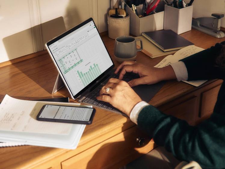 Someone working at a wooden desk with a laptop, notebooks and pots of pens