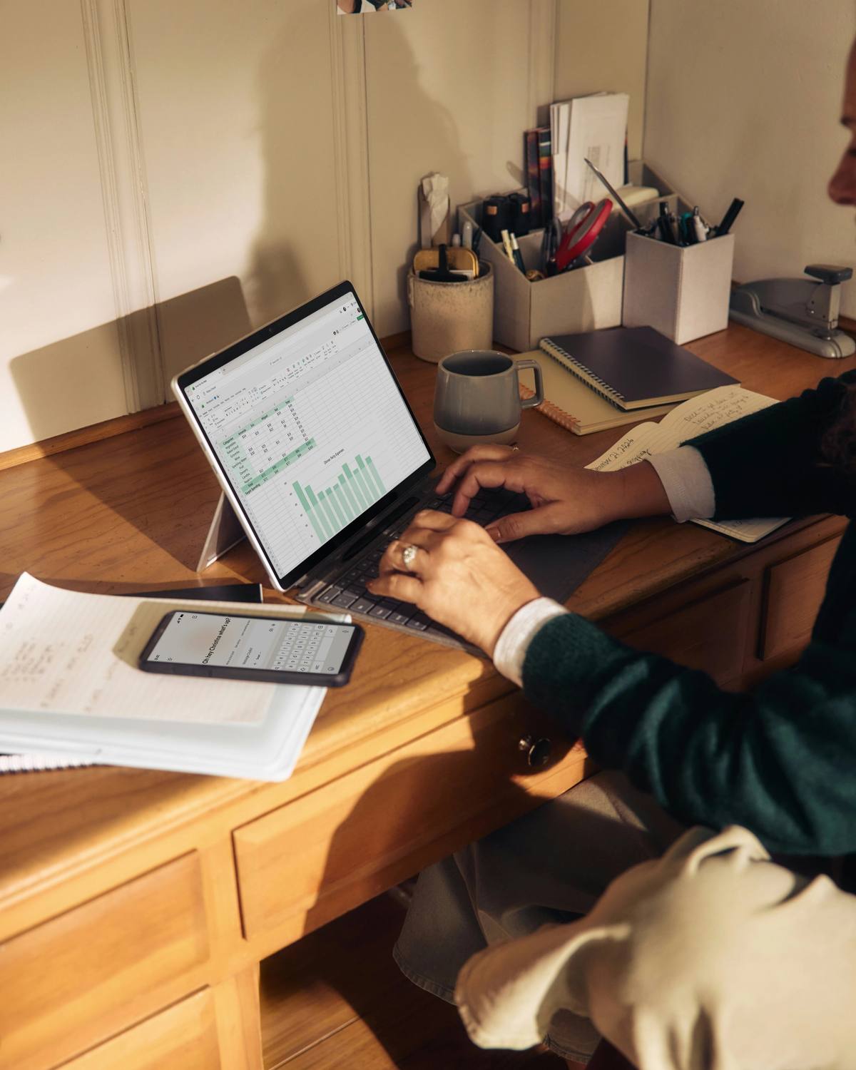 Someone working at a wooden desk with a laptop, notebooks and pots of pens
