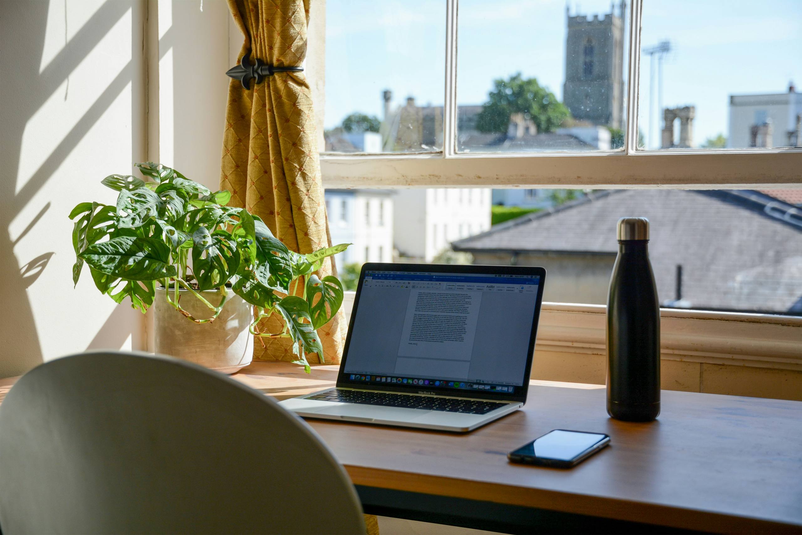 A desk by a sunlit window with a laptop, a plant and a view over rooftops