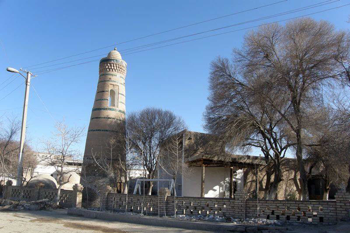 Das historische Minarett Abdal Bobo in der UNESCO-Altstadt von Chiwa, Usbekistan, umgeben von Bäumen und traditioneller Archi