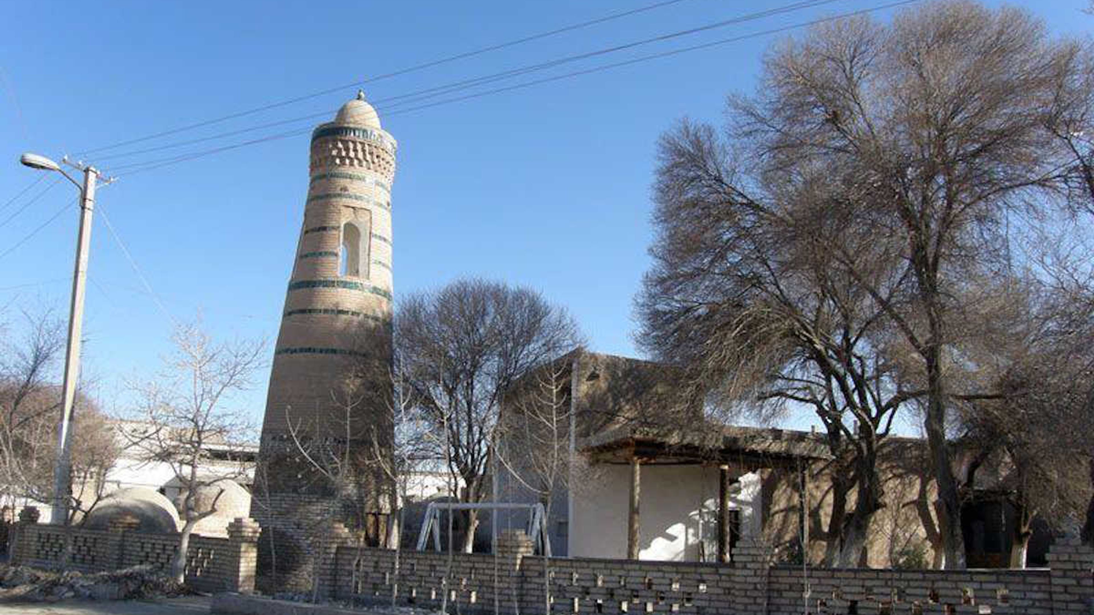Das historische Minarett Abdal Bobo in der UNESCO-Altstadt von Chiwa, Usbekistan, umgeben von Bäumen und traditioneller Archi