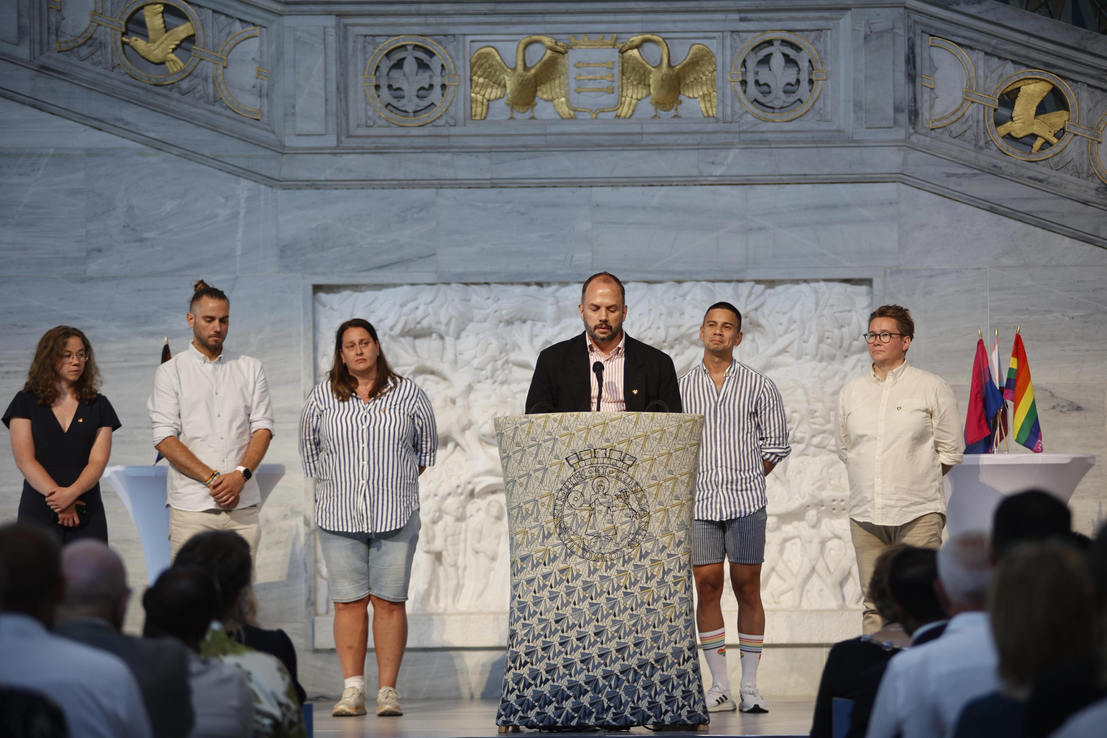 En mann står på talerstolen i Oslo rådhus. Bak ham står flere personer med alvorlig ansiktsuttrykk.