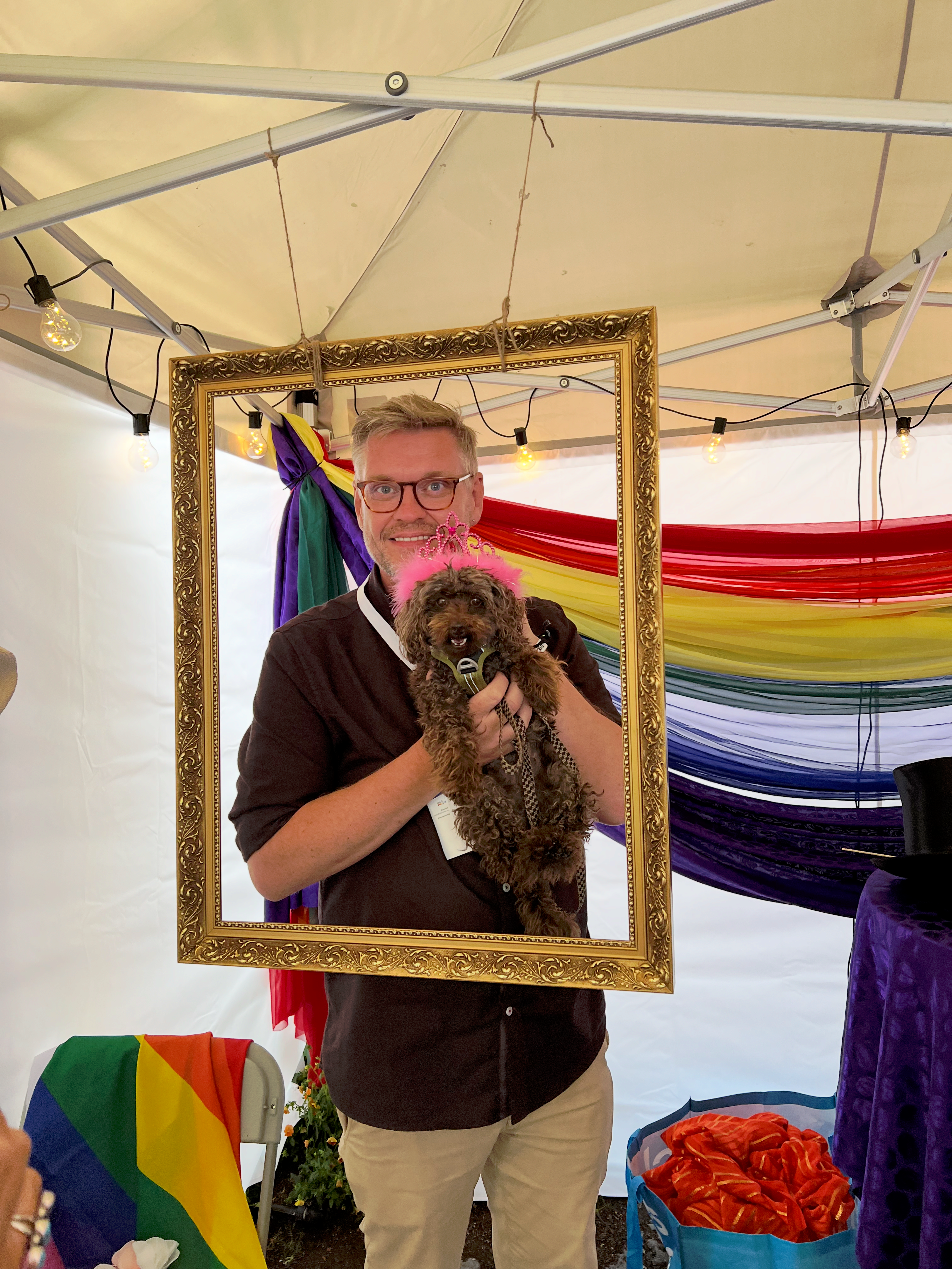 En mann holder opp en hund og smiler. De står inni en bilderamme med regnbuebanner i bakgrunnen. Hunden har rosa tiara på hodet.