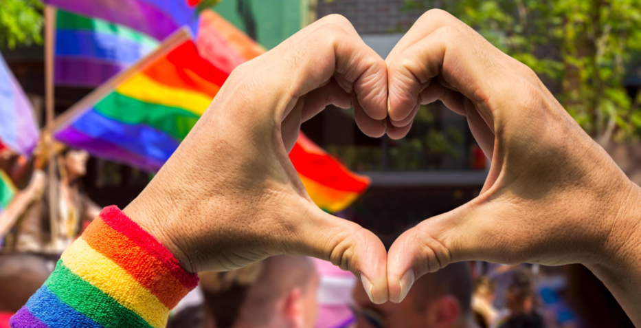 To hender lager hjertesymbol. Personen har på seg pulsvarmer i regnbuefarger, og i bakgrunnen ses flere regnbueflagg.