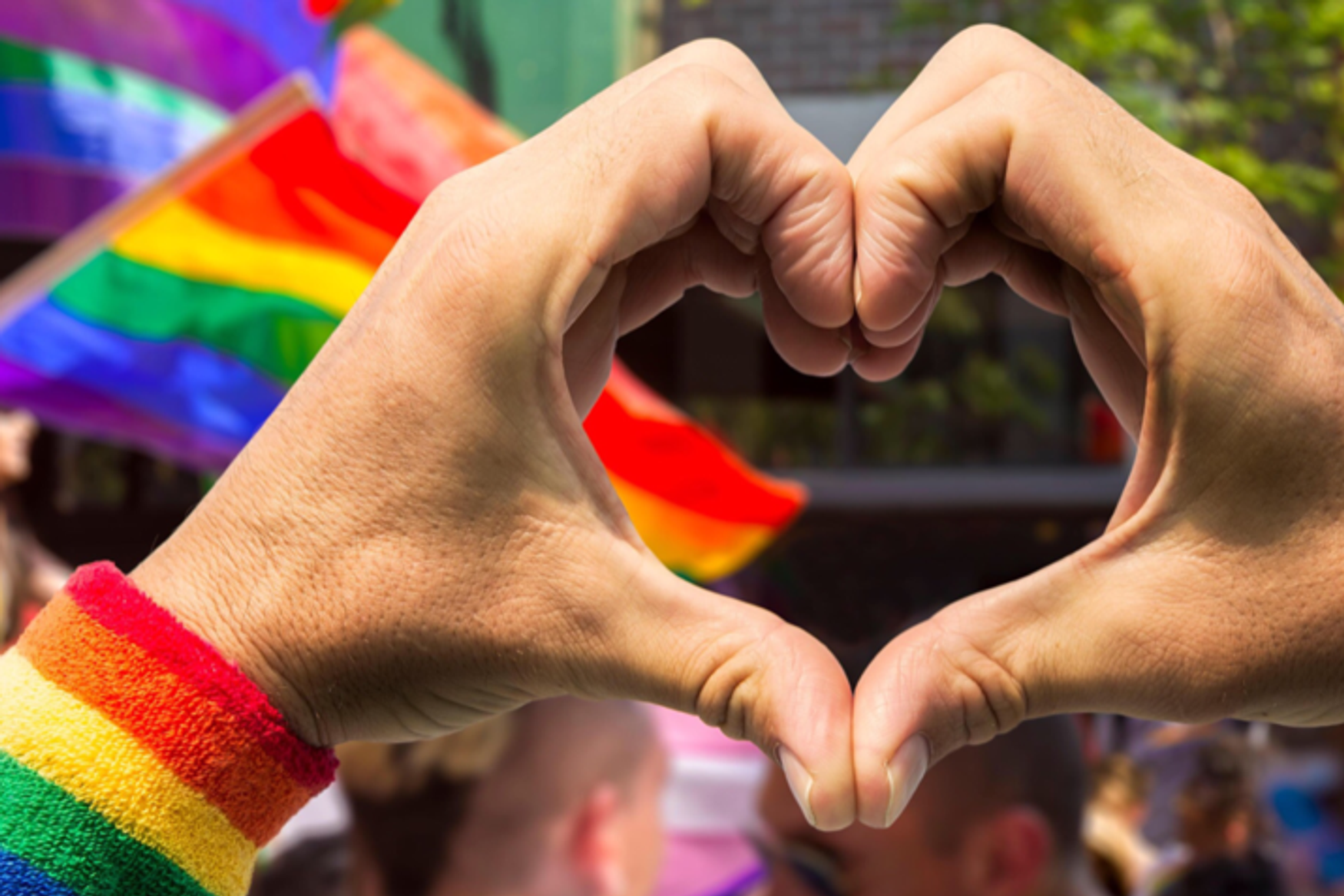 To hender lager hjertesymbol. Personen har på seg pulsvarmer i regnbuefarger, og i bakgrunnen ses flere regnbueflagg.