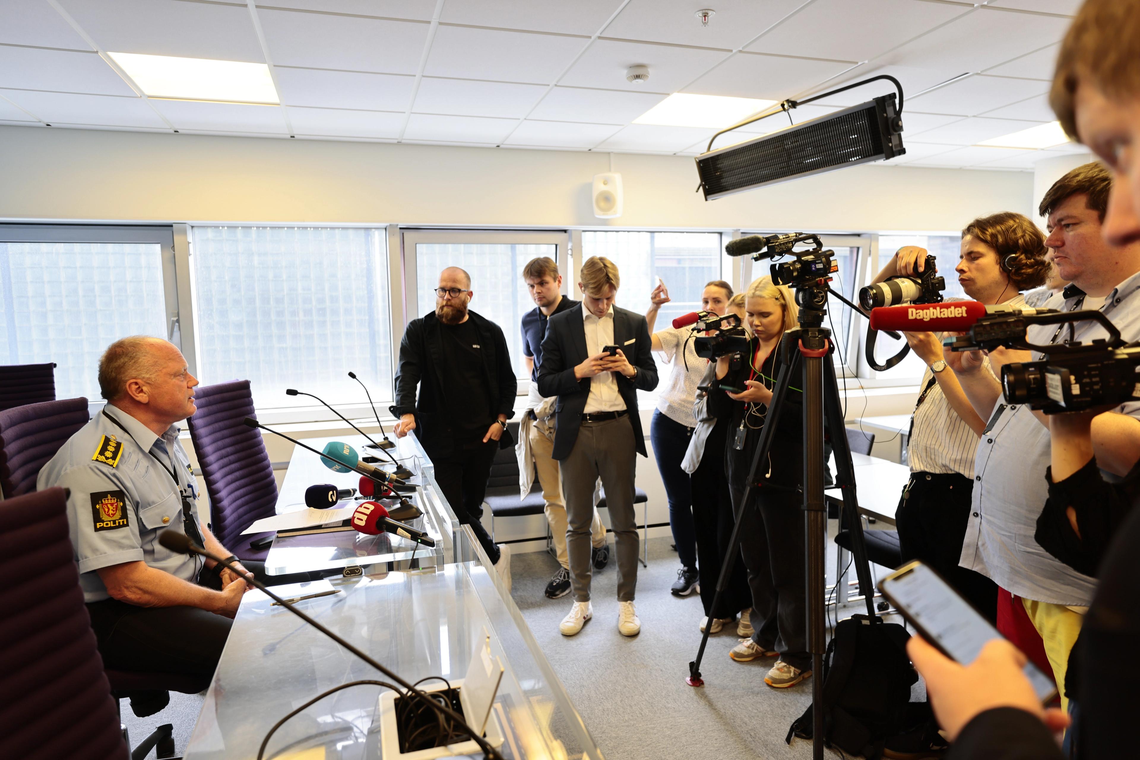 En politimann sitter bak et bord. På andre siden står mange journalister med kameraer og mikrofoner.