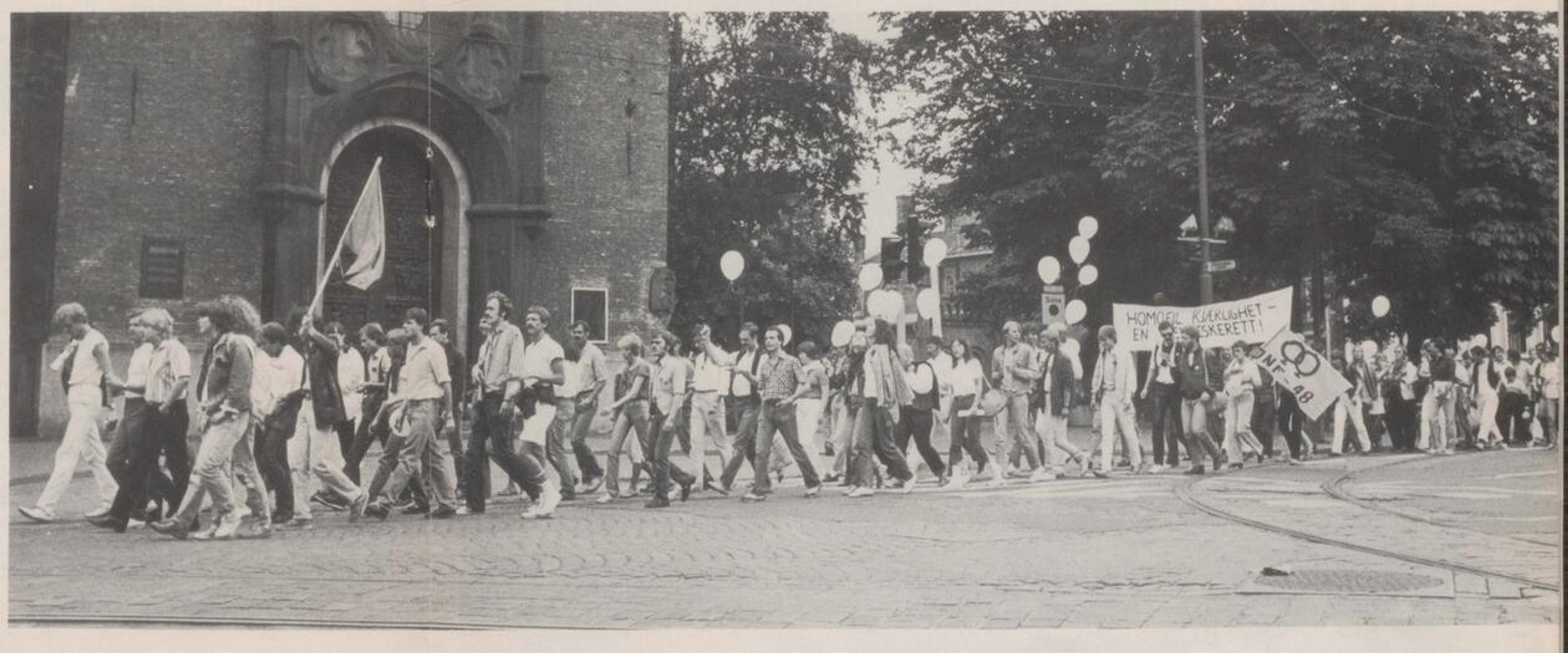 Sort-hvitt bilde av et opptog i Oslo. Deltakerne bærer bannere med slagord for homofil frigjøring.