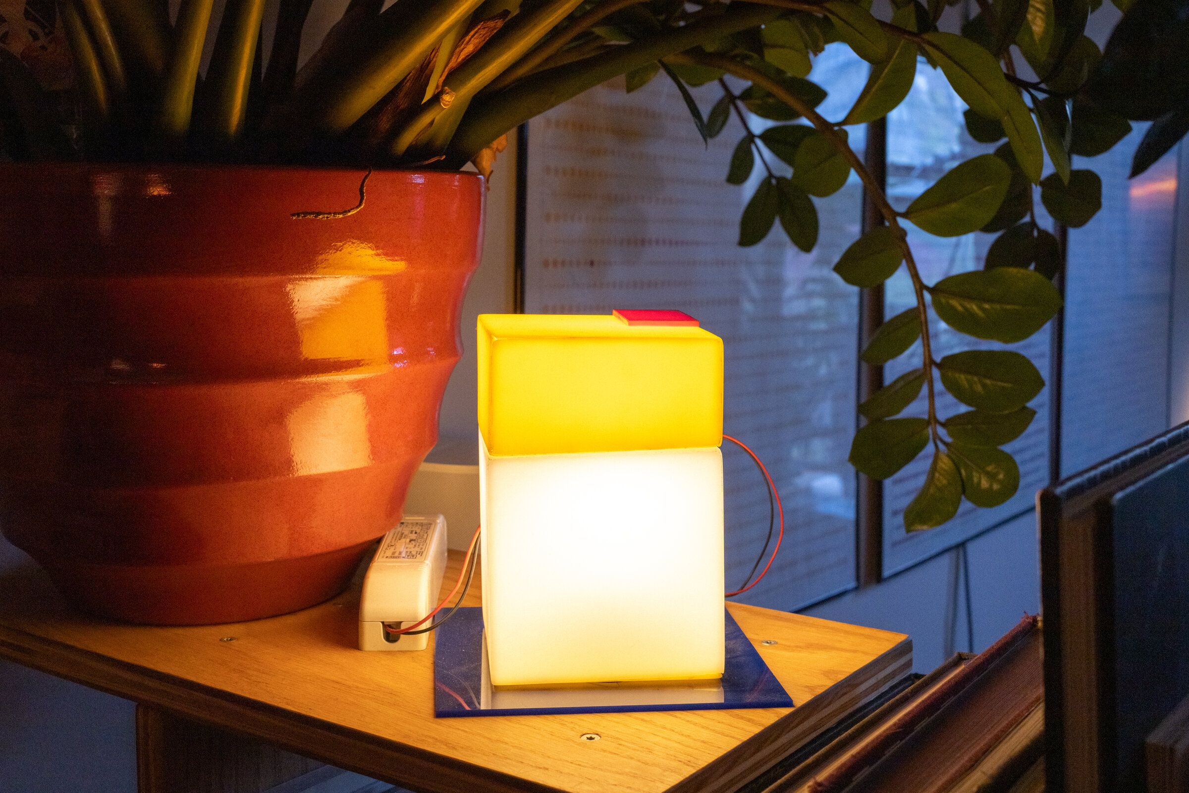 Parts and all on a tabletop next to a terra-cotta planter: a glowing white cube with a yellow cap and a small red square on top, blue plate at the base