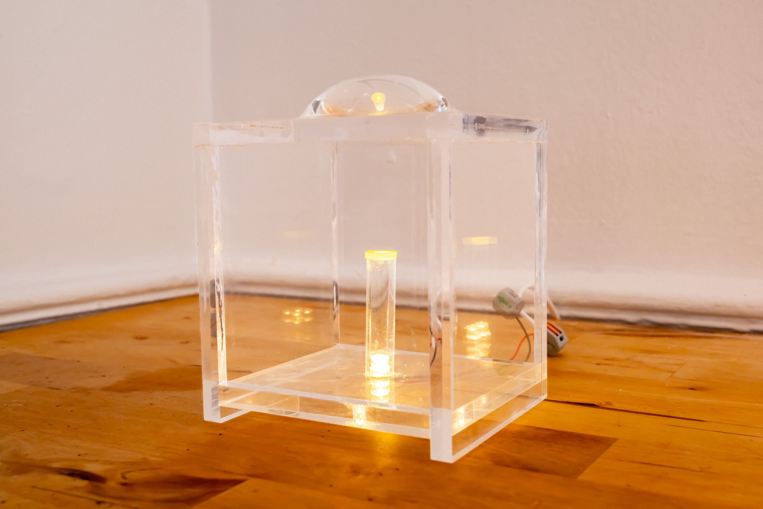 Three-quarter view of internal artifact: clear acrylic cube on a wooden floor, lens dome on top, lit vial visible inside, leds glowing around the base