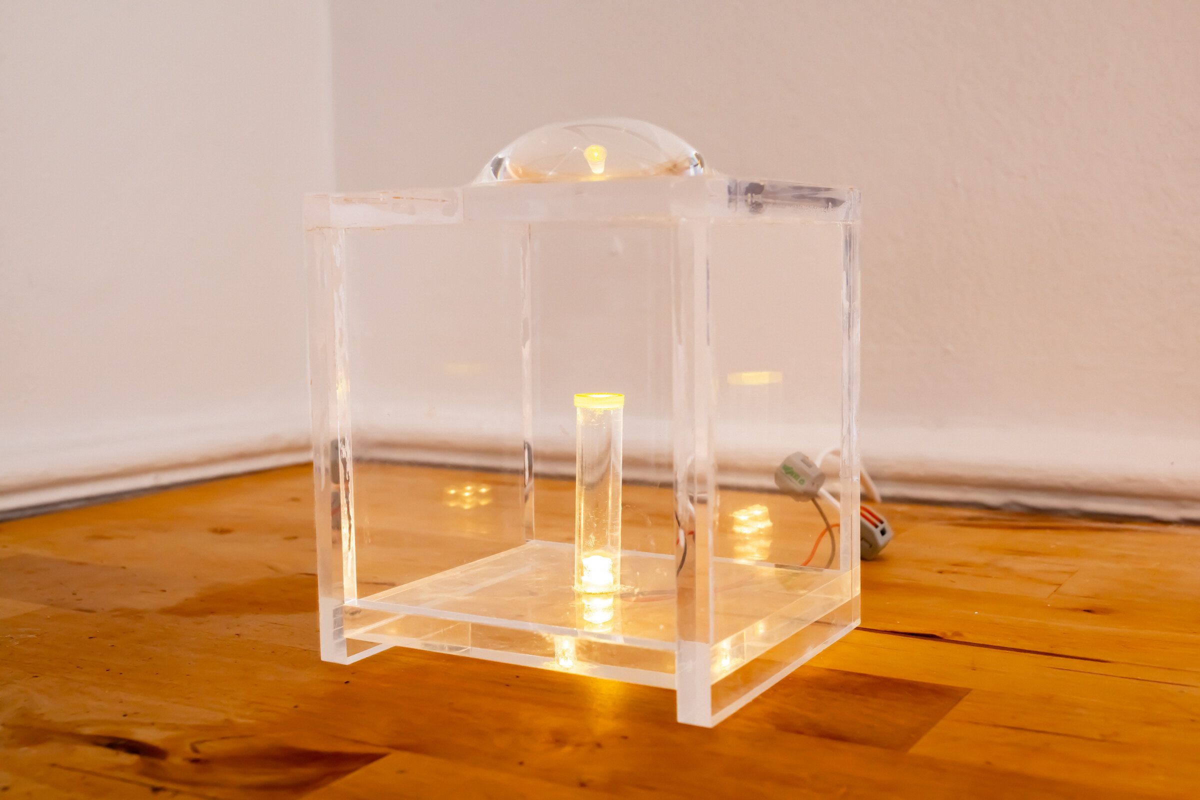 Three-quarter view of internal artifact: clear acrylic cube on a wooden floor, lens dome on top, lit vial visible inside, leds glowing around the base