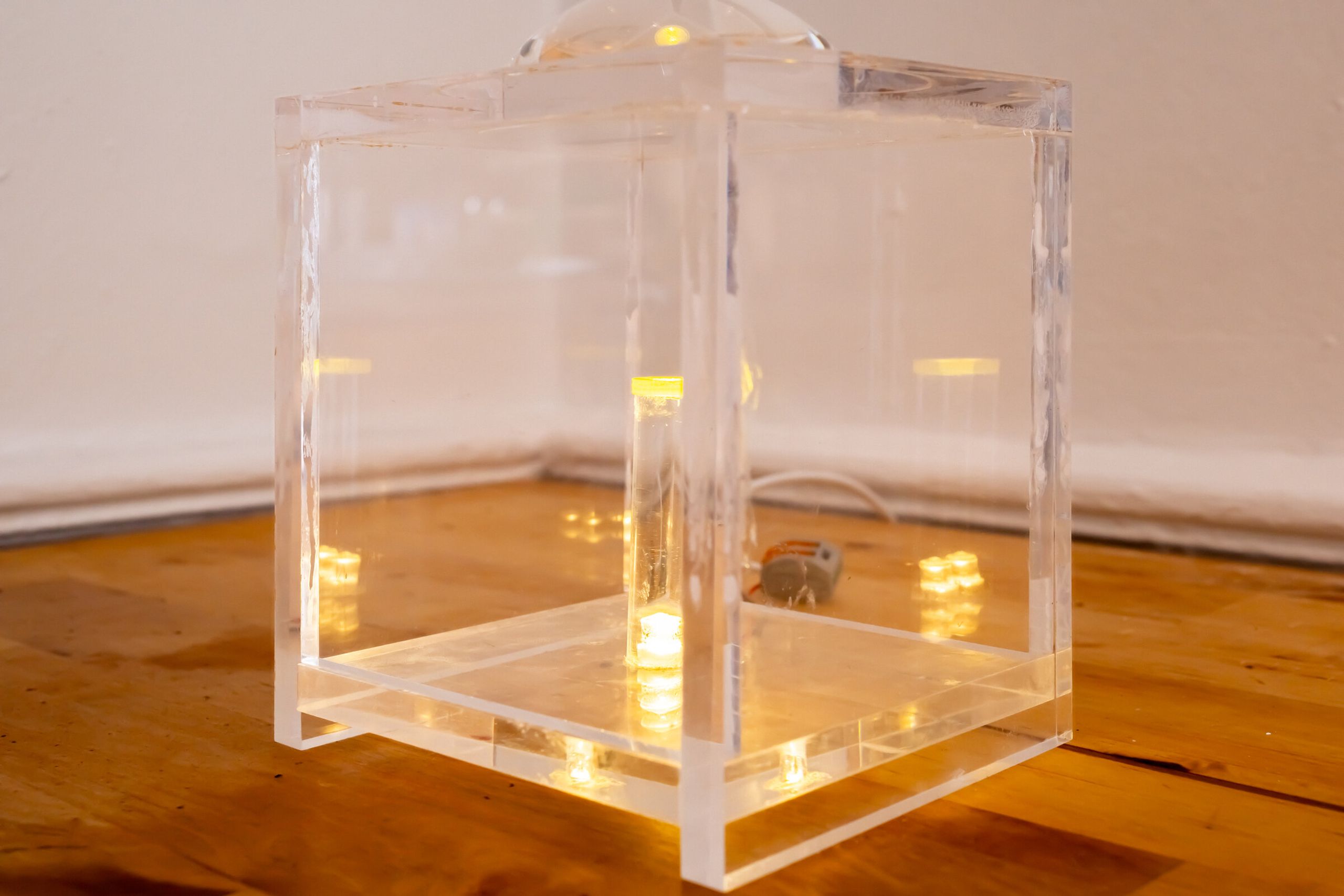 Off-angle view of internal artifact: the clear cube on the wooden floor seen from above, lens visible at top edge, lit elements glowing inside