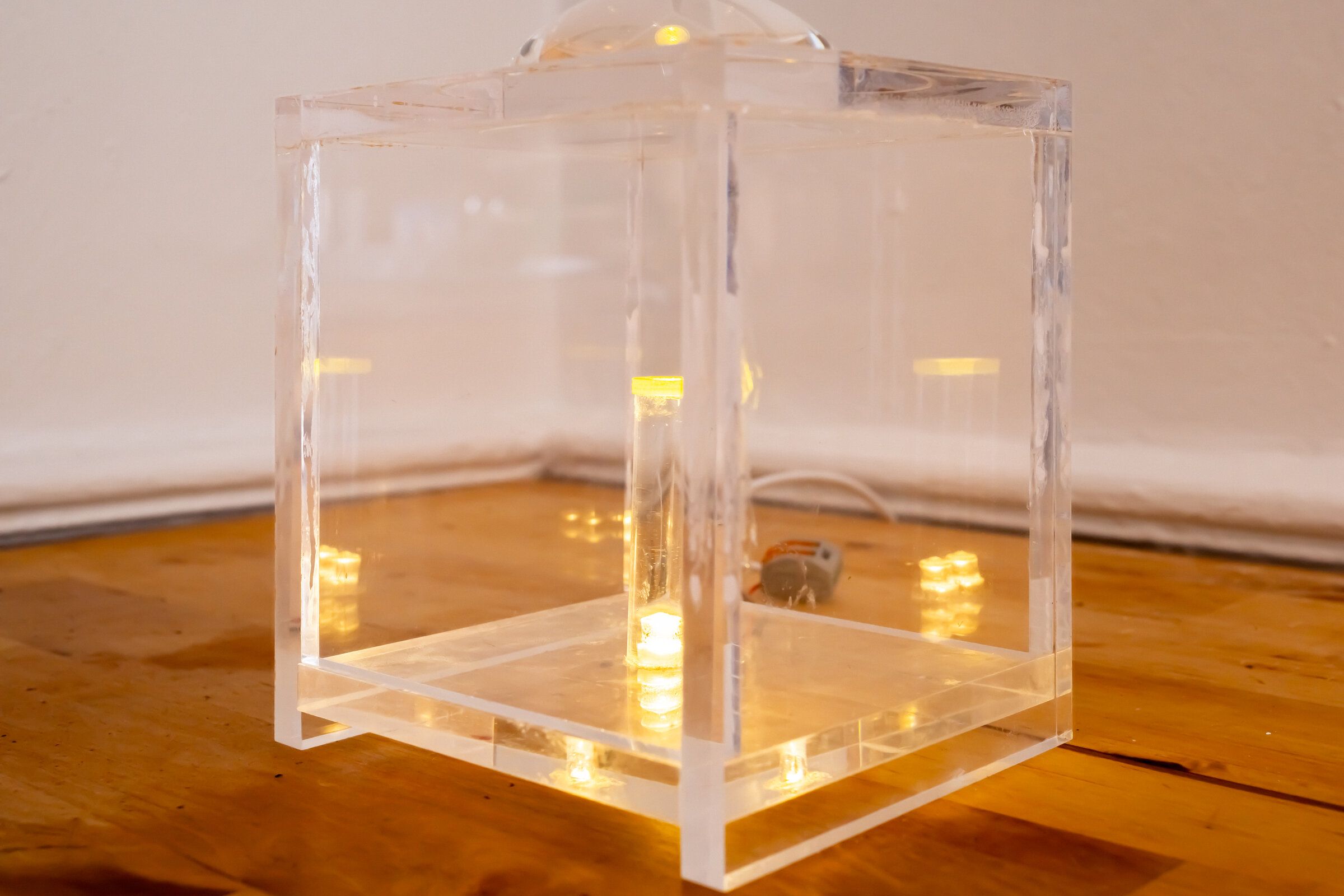 Off-angle view of internal artifact: the clear cube on the wooden floor seen from above, lens visible at top edge, lit elements glowing inside
