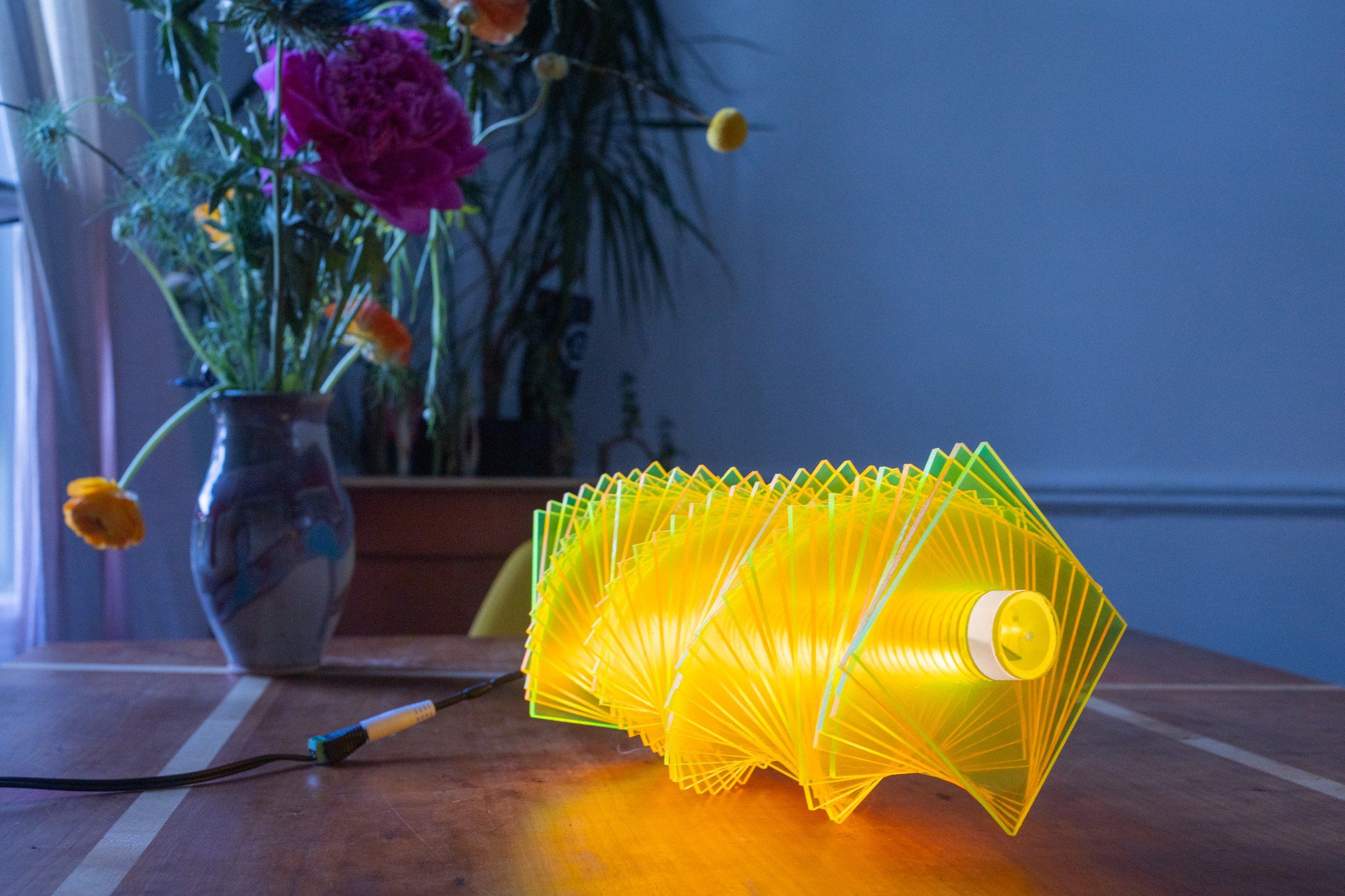 Radiator IV on a wooden tabletop: green and yellow fluorescent plates spiraling around an LED tube, a vase of cut flowers and a curtain visible beyond, the cord running off to the left
