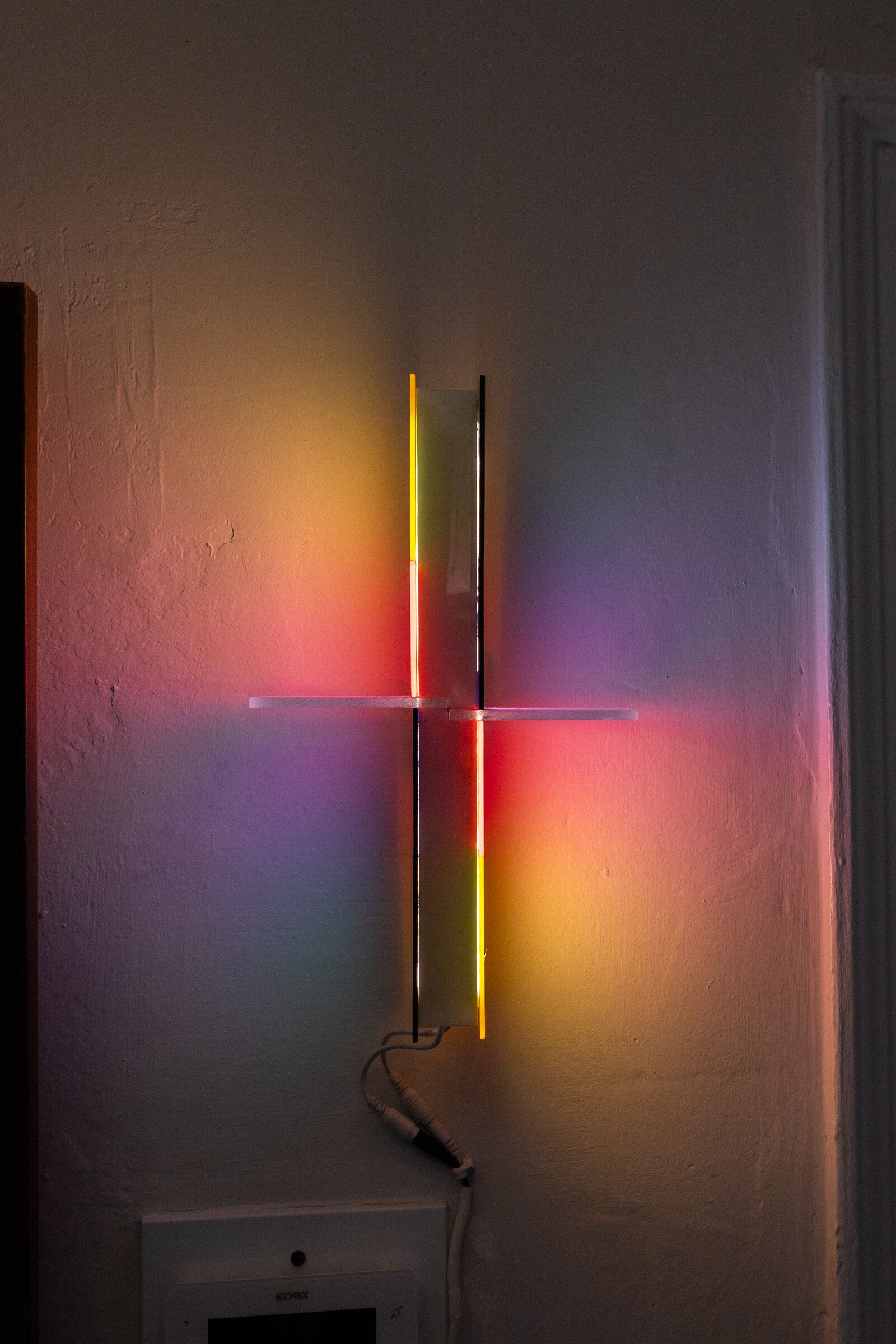 Irreversible: a wall-mounted cross of light with colored halos washing the wall around it; yellow and orange above the cross arms, red and pink below