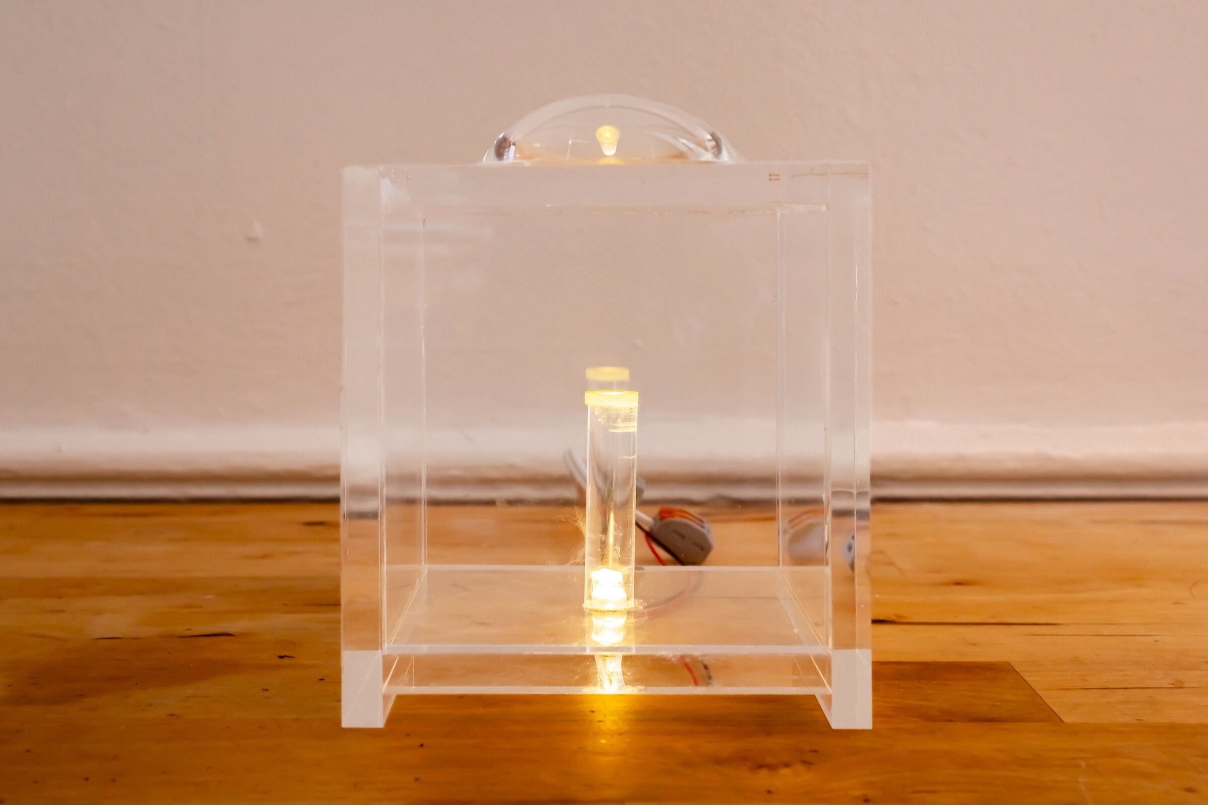 Internal artifact on a wooden floor: a small clear acrylic cube containing a lit cylindrical vial at its center, a clear lens dome resting on top