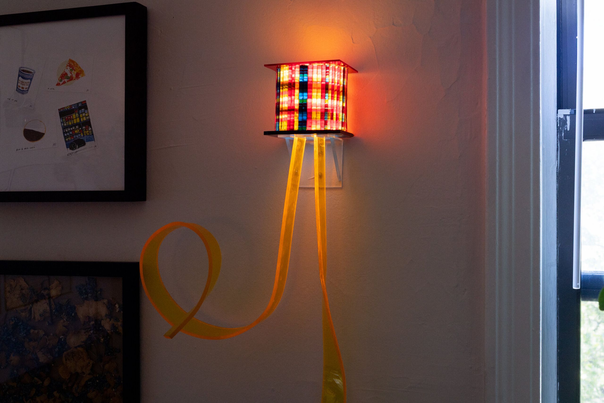 Centered view of i've been thinking about flowers: the warm-lit box mounted on the wall, an orange tendril curving outward below in a loop