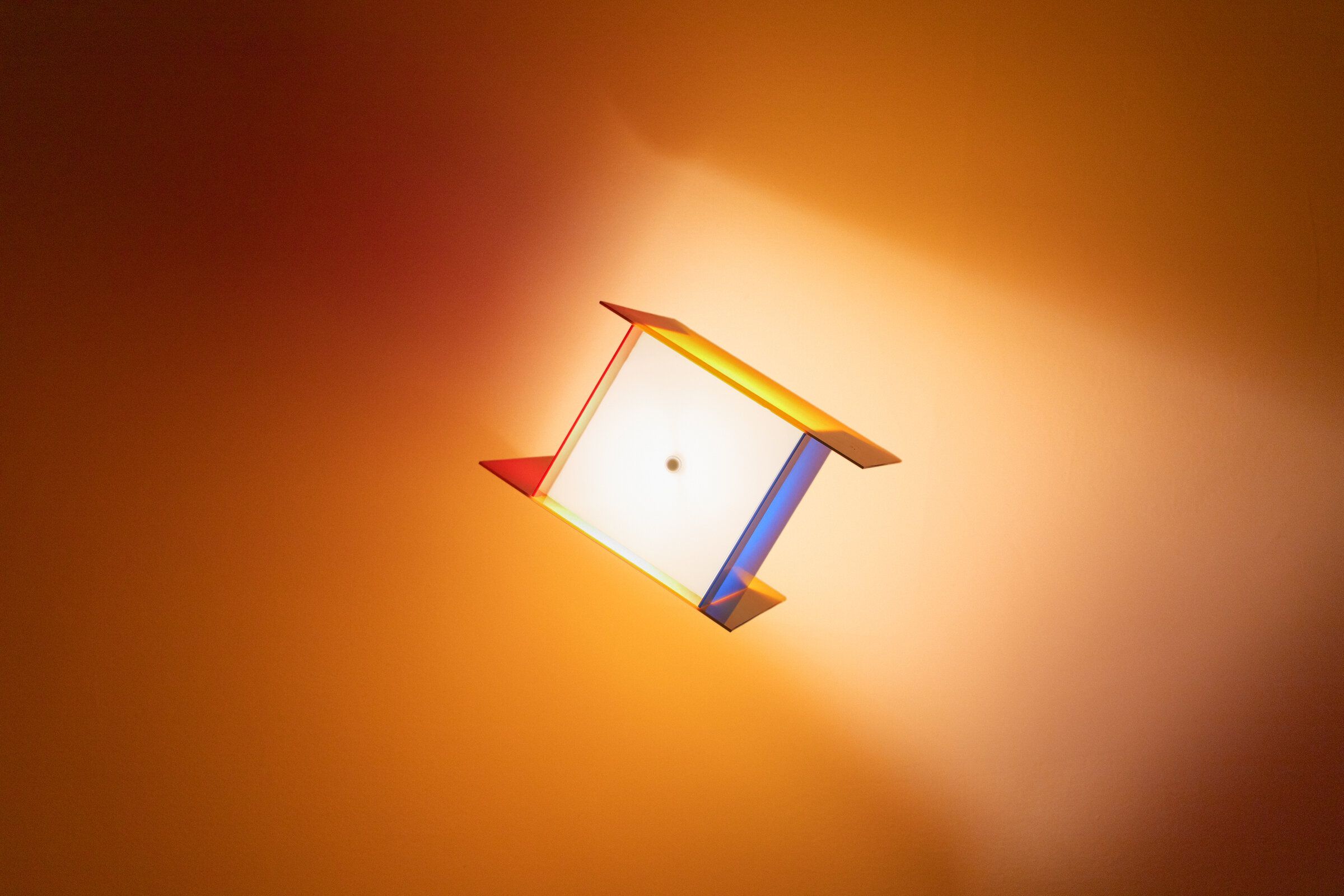 Square one mounted on a ceiling: a small angled square of yellow acrylic throwing warm splashes of color onto the ceiling around.