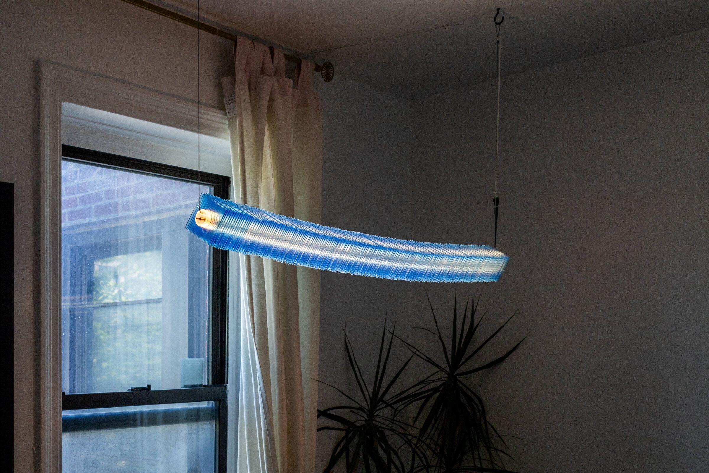 Radiator I hung horizontally in a room corner: a long bar of stacked blue acrylic plates, the warm filament glowing through the interior at one end, suspended from the ceiling by two cables