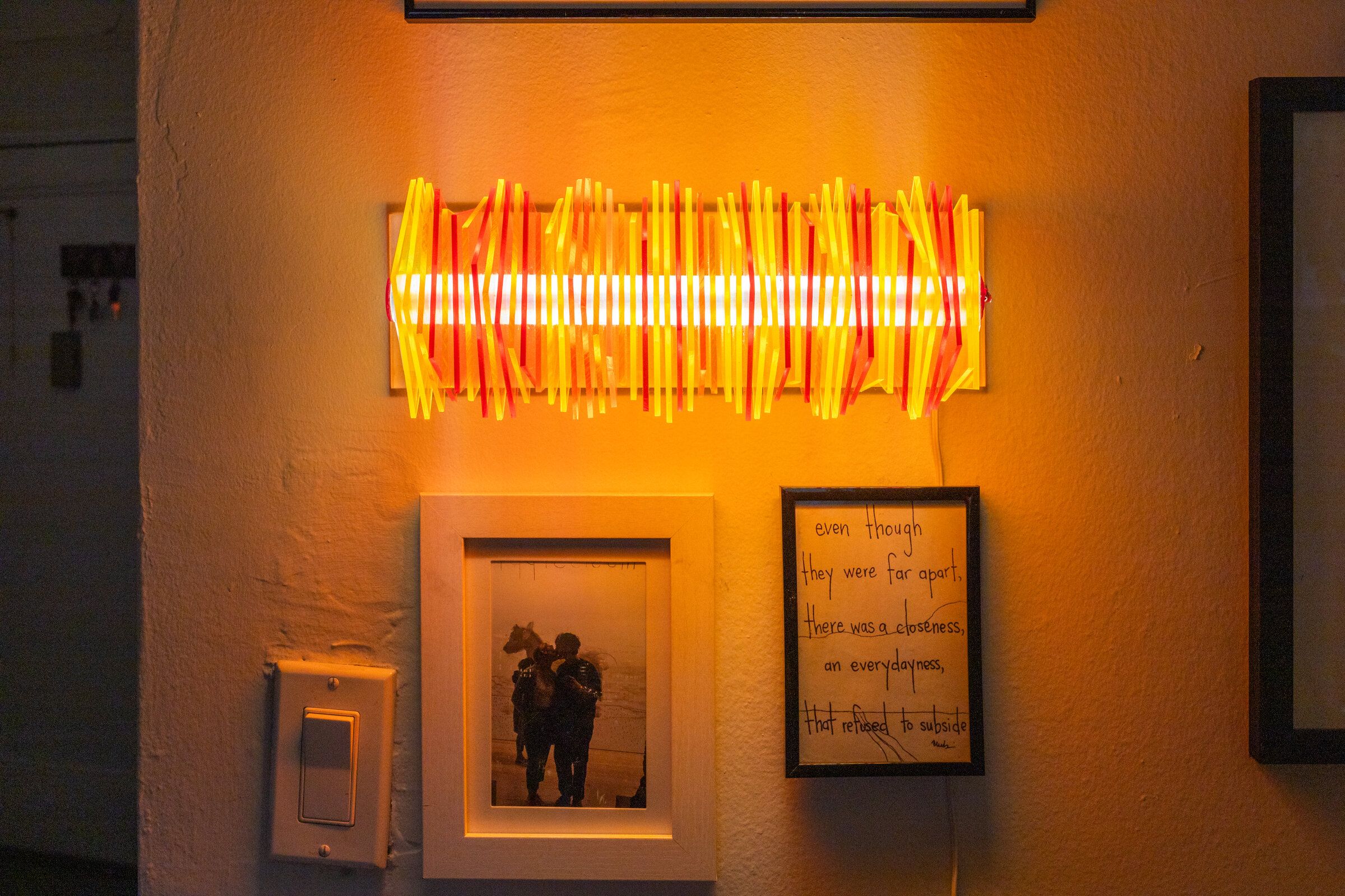 Radiator II mounted on a wall: a horizontal bar of warm yellow, orange, and red acrylic plates, the warm filament glowing through the interior, surrounded by framed art and a light switch