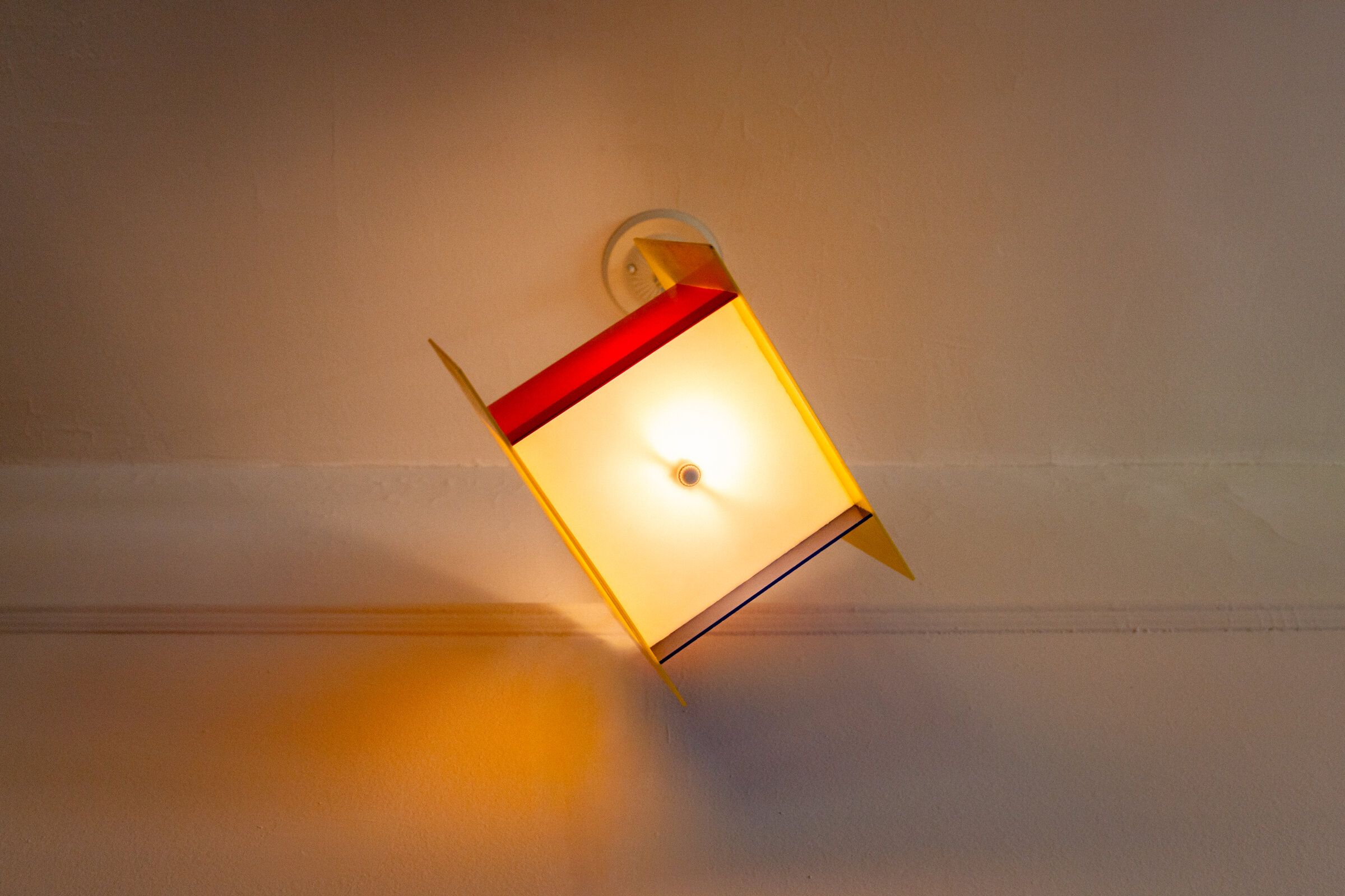 Square one mounted at an angle on a wall, frosted front lit from within, warm yellow light spreading down the wall