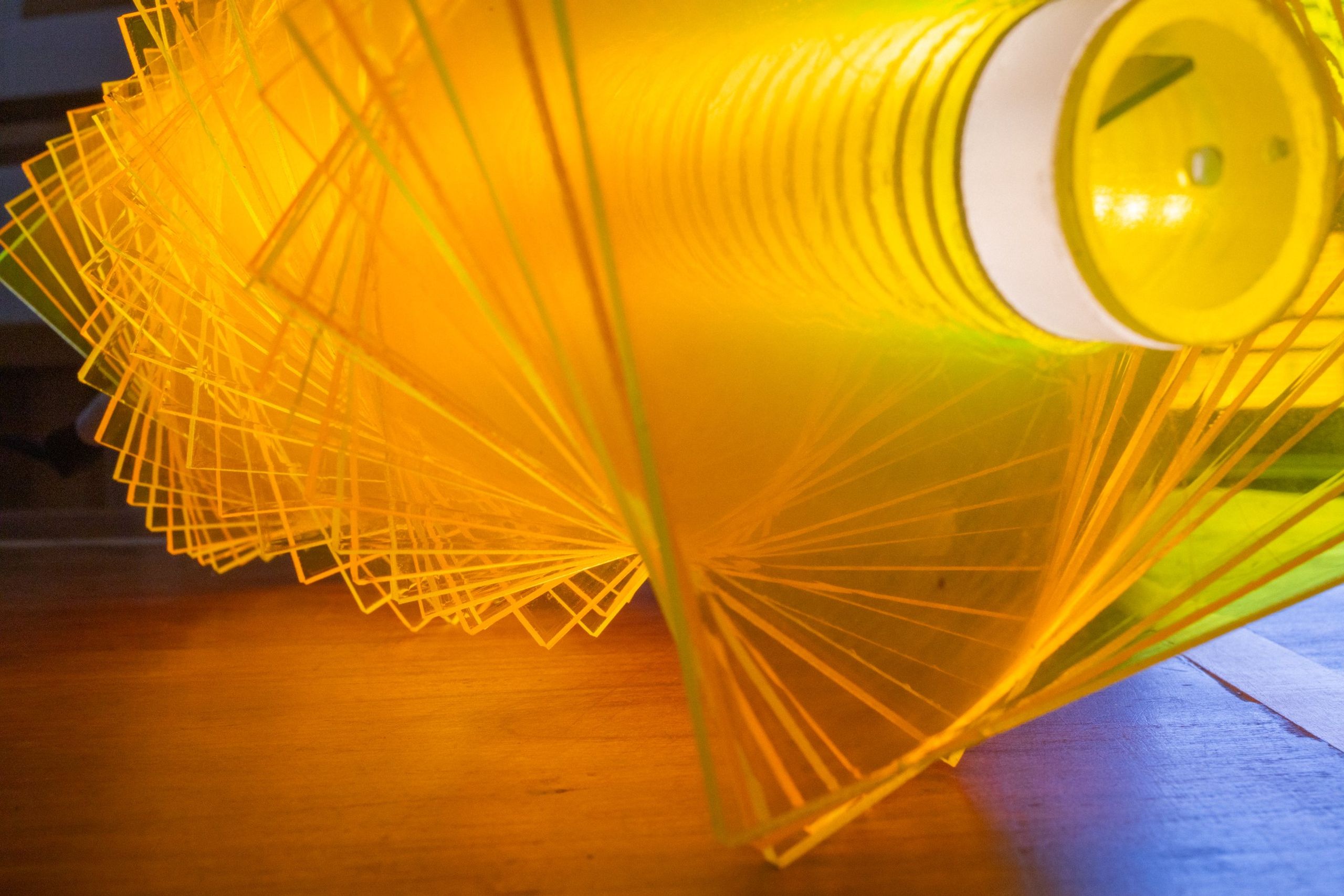 Three-quarter view of radiator IV from above: green and yellow fluorescent acrylic plates fanned around a horizontal LED tube, the ceramic socket visible at the right end, plates casting warm light onto the wood surface below