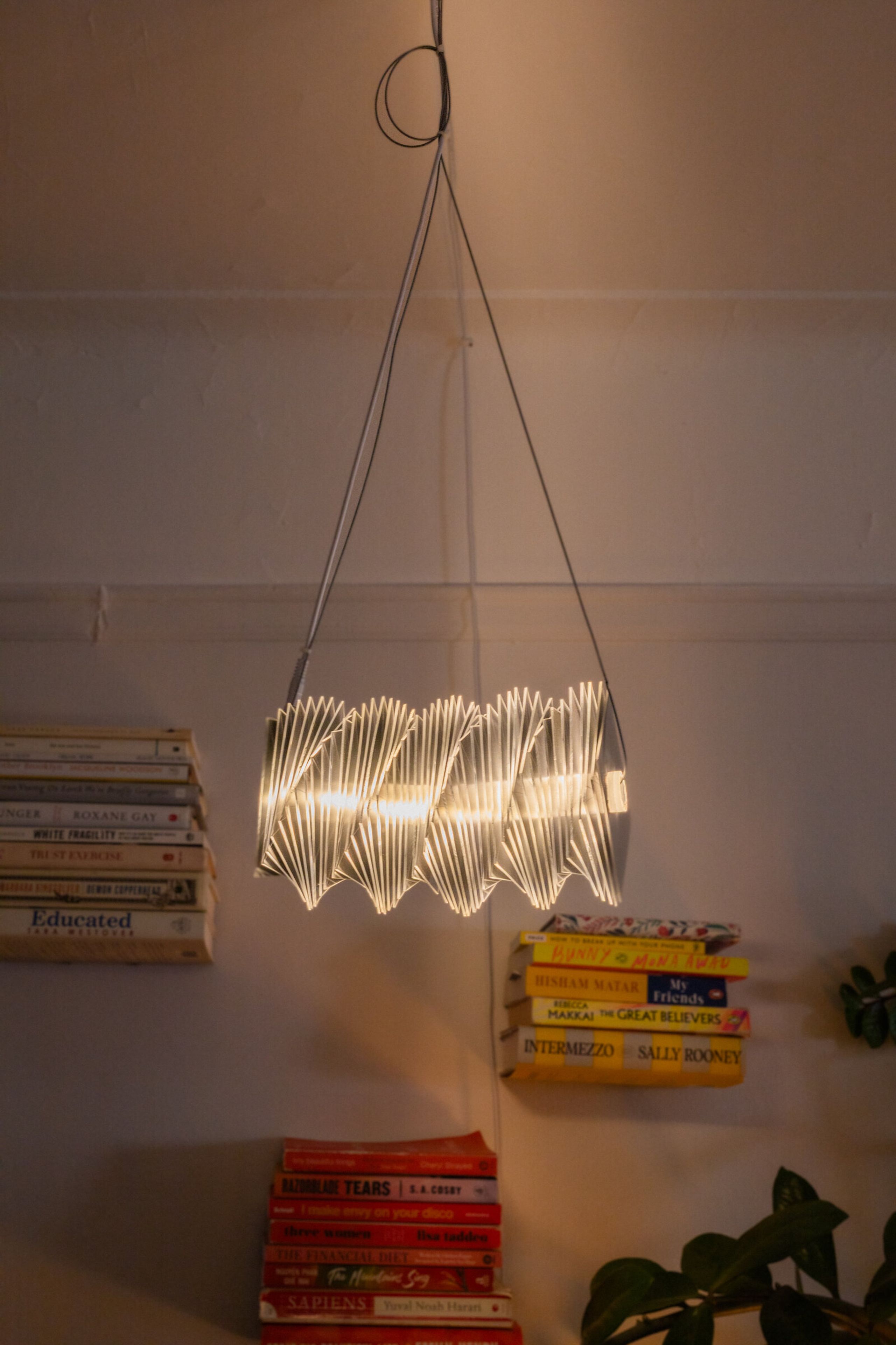 Radiator III hung from the ceiling in a living room: a horizontal cylindrical form glowing warm at its center, mirrored plates radiating outward, suspended above stacks of books