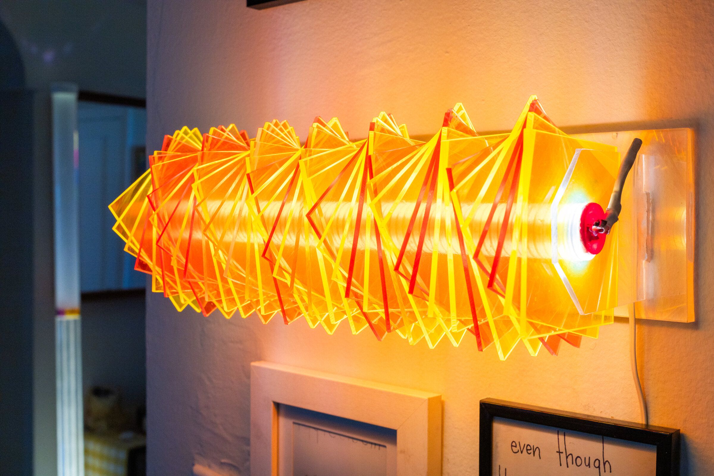 Side view of radiator II: stacked acrylic plates in yellow, orange, and red rotated around a warm linear core, mounted to the wall via a clear acrylic bracket, ceramic socket visible on the right end