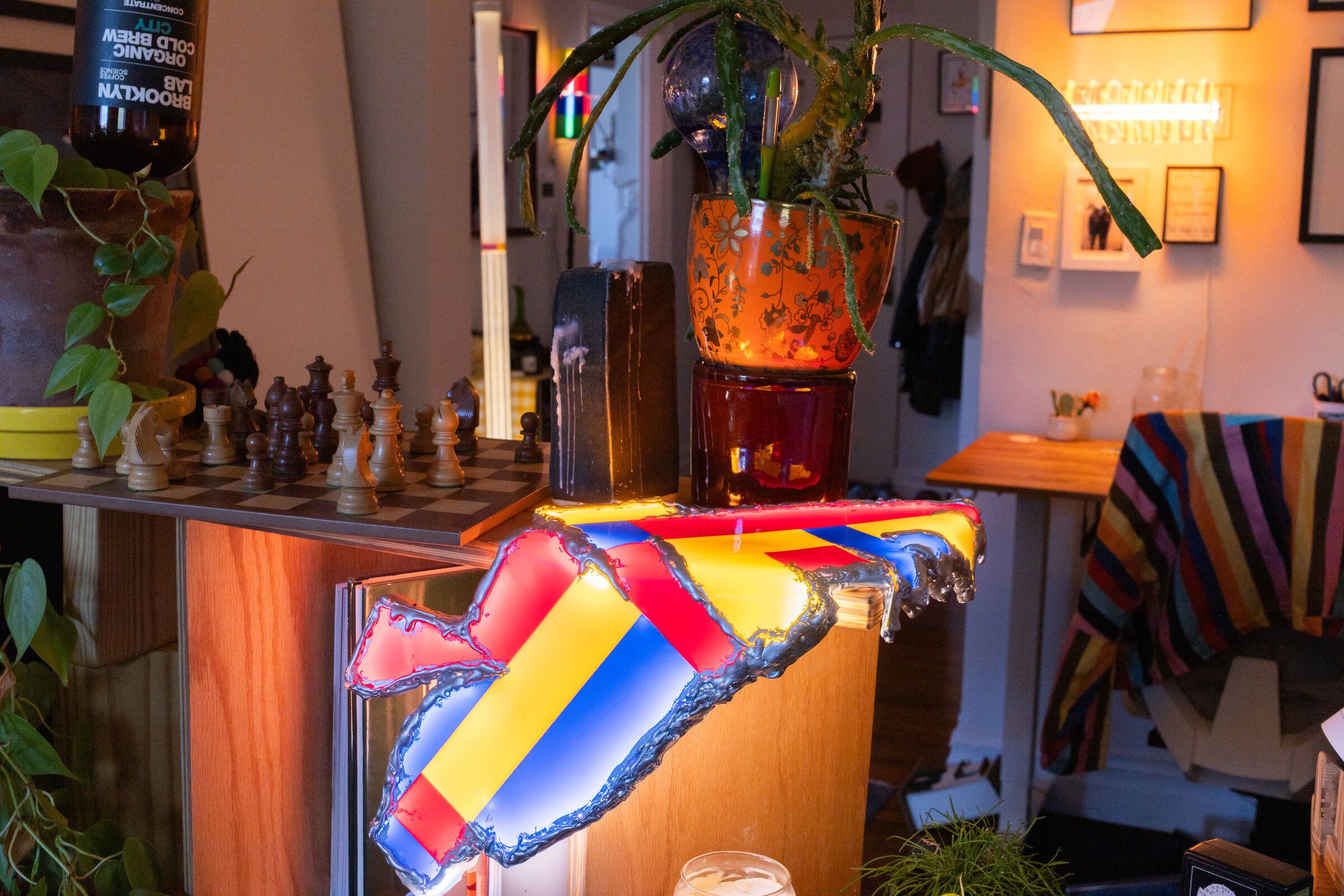 Shelf life draped over the corner of a wooden shelf next to a plant and a chess board: a folded mosaic of red, yellow, and blue panels with molten silver-painted edges, glowing