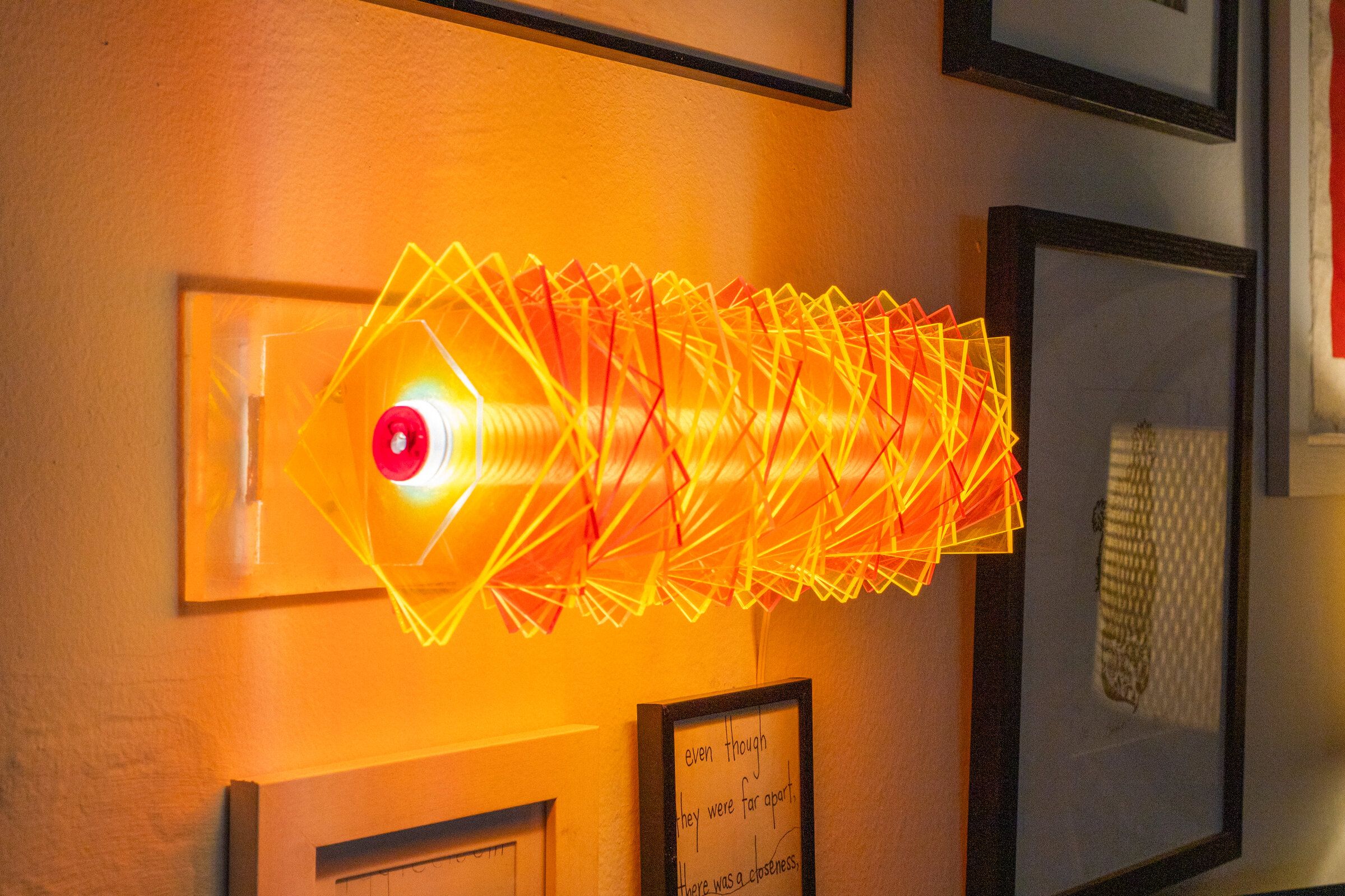 Three-quarter view of radiator II from above: the warm bulb visible at the left end, plates rotating outward in red and yellow and orange, casting a warm halo on the wall behind