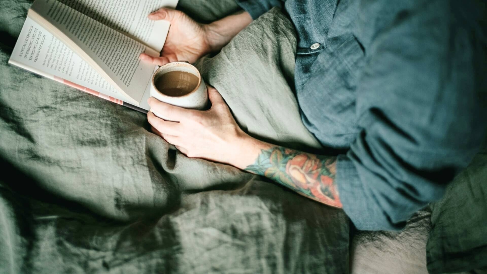 Person with tattooed arm holding a cup of coffee and reading a book in bed with grey-green bedding