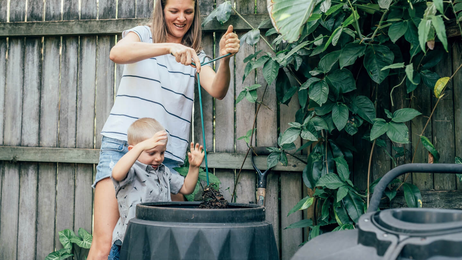 A woman and a young child are engaged in composting activities with a black compost bin in a home garden setting