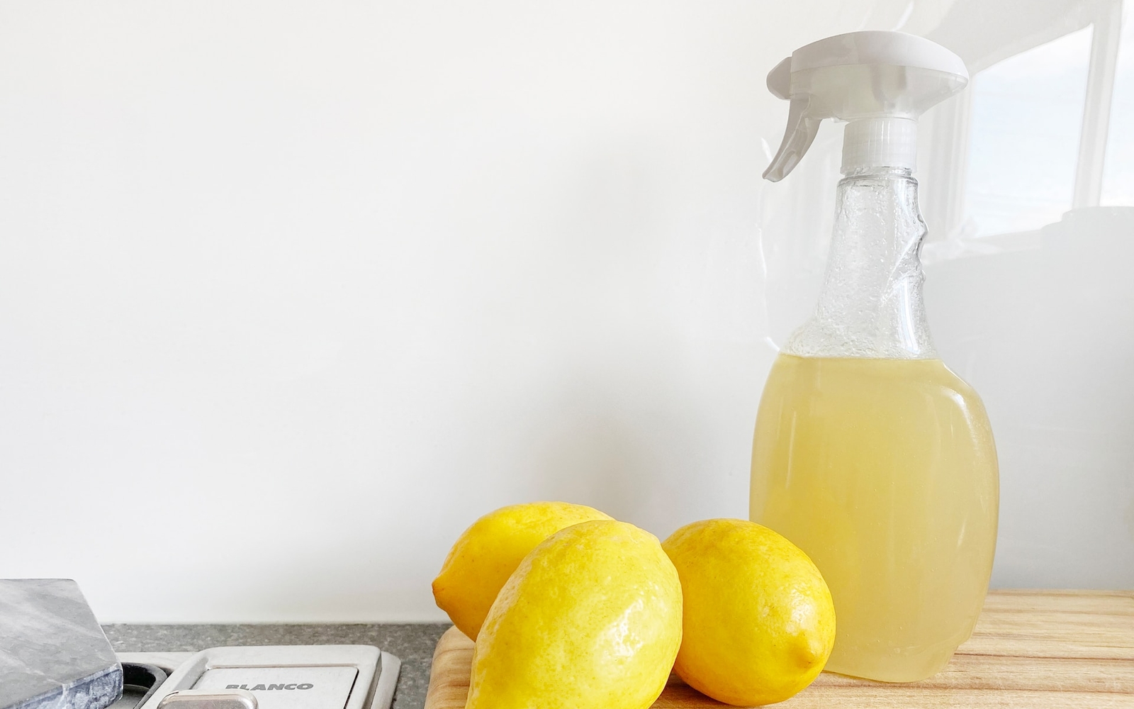 A homemade cleaning solution in a clear spray bottle next to two whole lemons on a wooden kitchen counter.