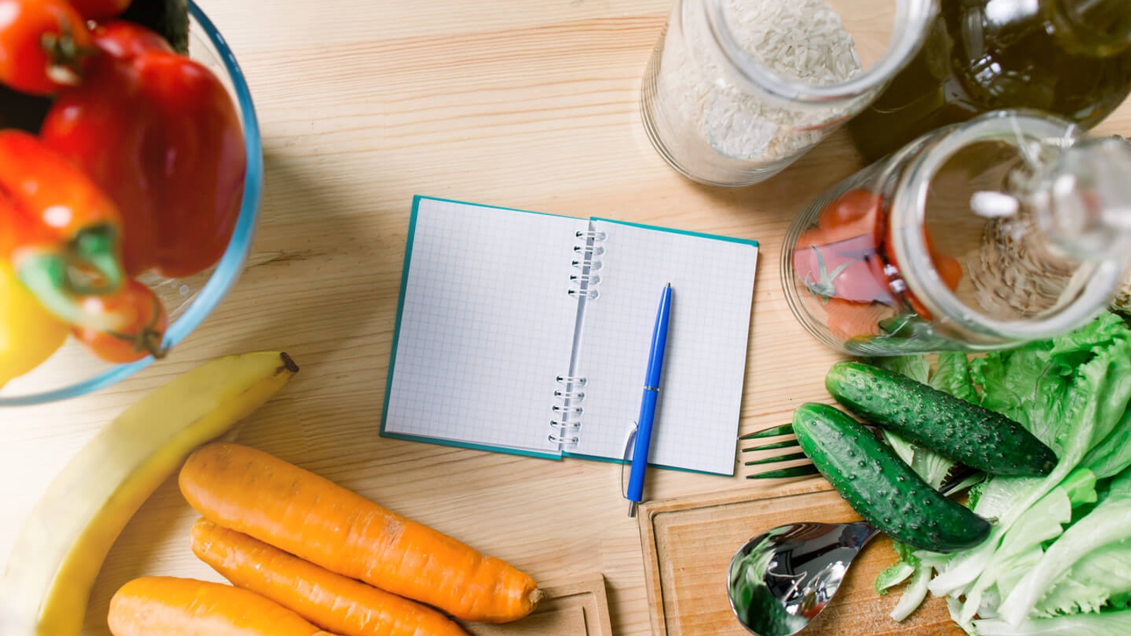 A kitchen countertop with a variety of fresh vegetables, including carrots and bell peppers, and kitchen essentials like a notebook, a pen, and rice grains, suggesting meal planning or recipe preparation.