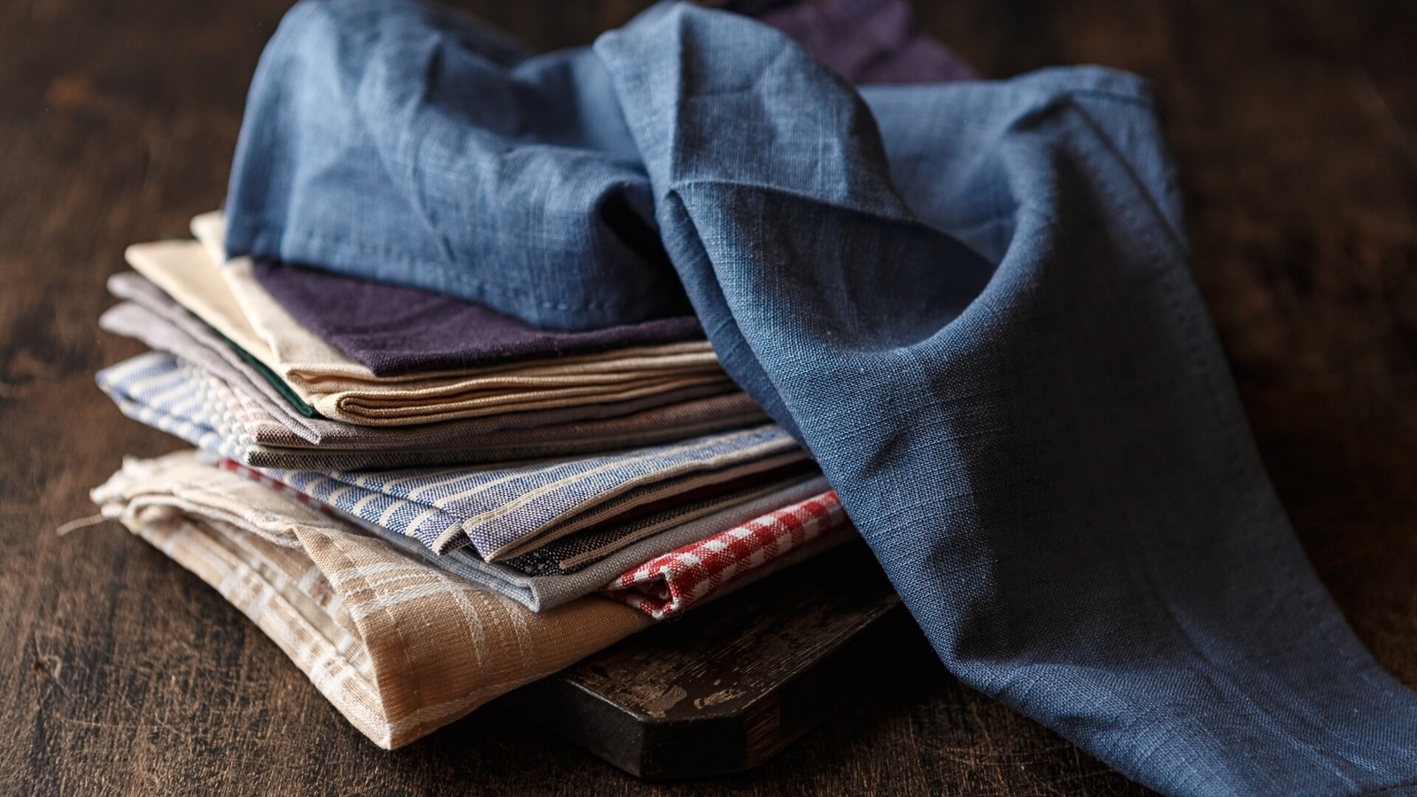 A stack of folded, multi-colored fabrics, including shades of blue, beige, and checkered patterns, on a wooden surface.