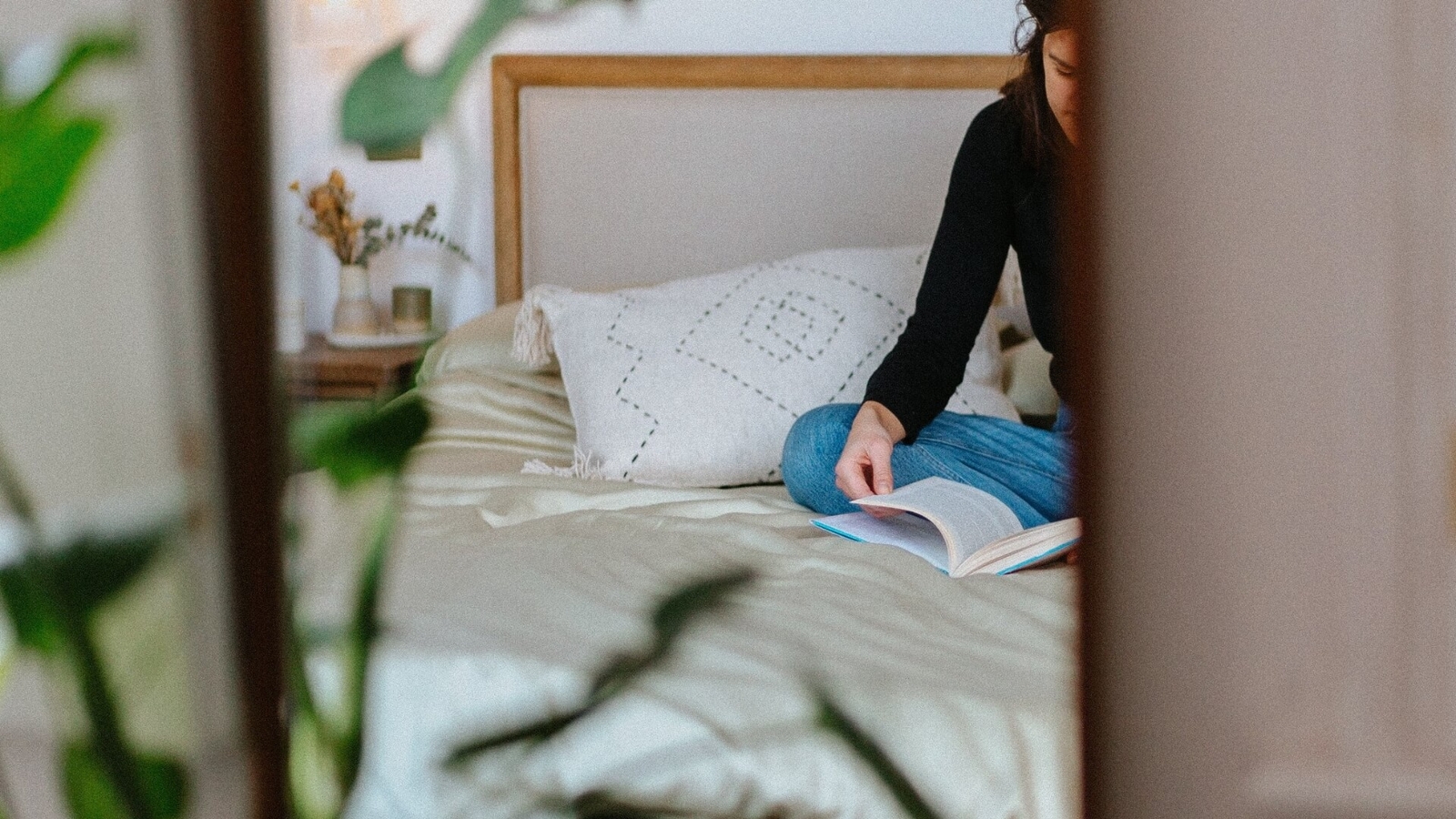 Person sitting on a bed with light-colored sheets, reading a book with plants in the foreground.