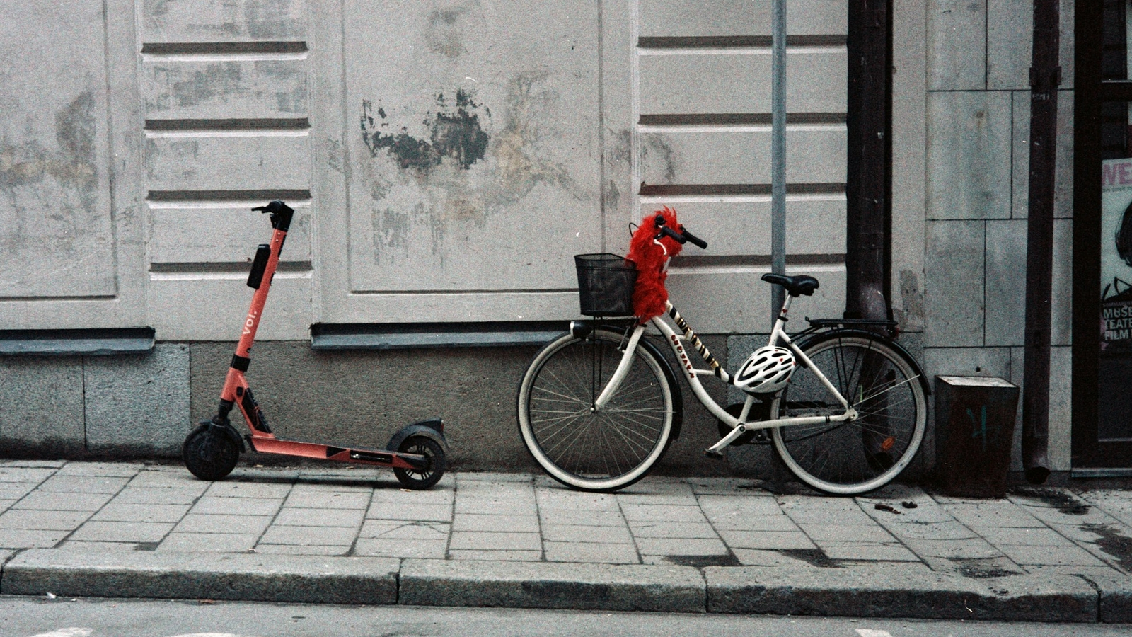 A red electric scooter and a white bicycle with a basket and a red feathered decoration parked side by side on a city sidewalk.