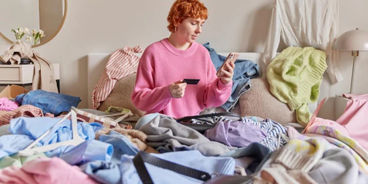 A person sitting on their bed surrounded by piles of clothes, looking at their phone
