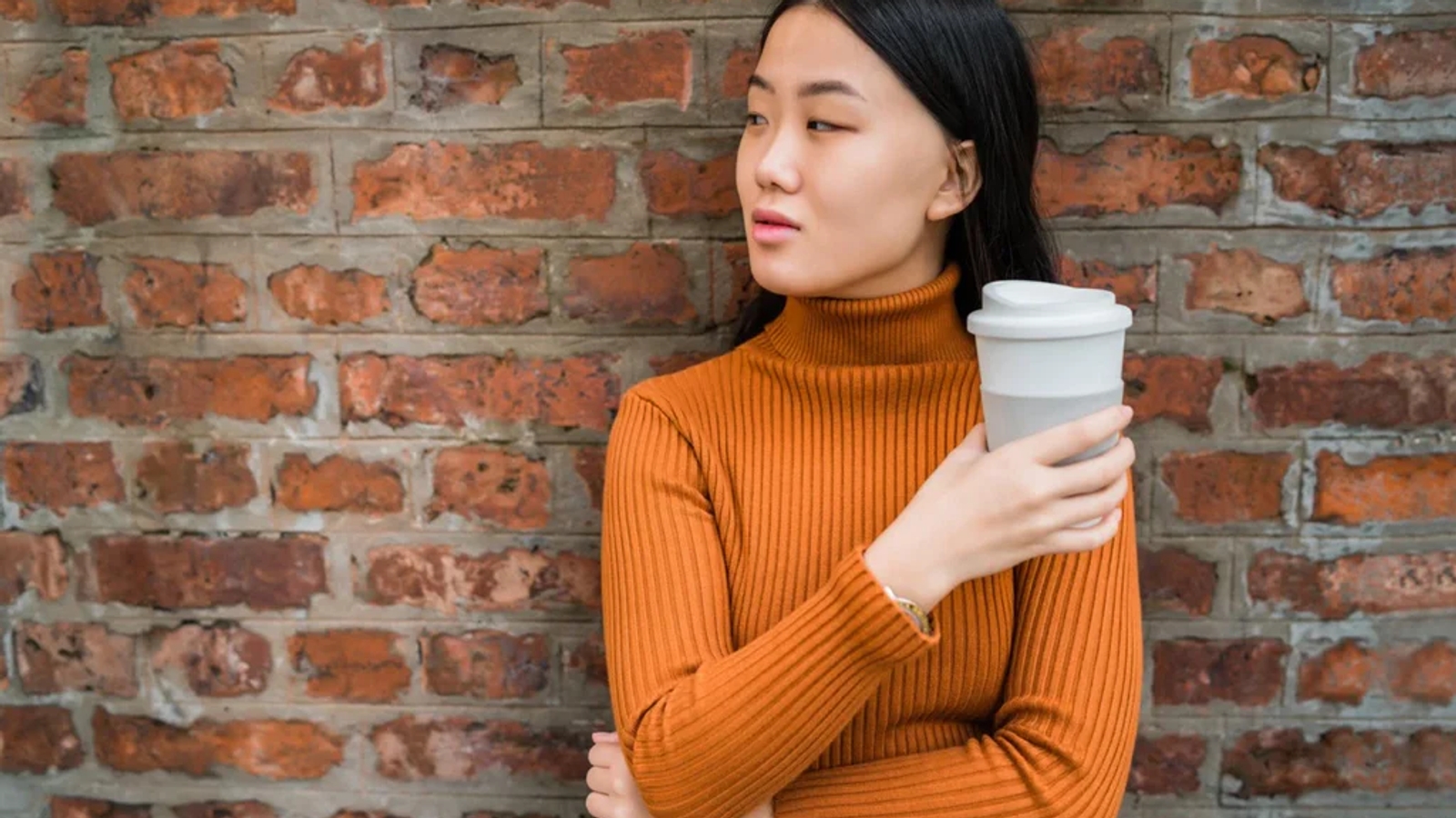 a woman standing with her arms crossed wearing an orange turtleneck and holding a reusable coffee cup