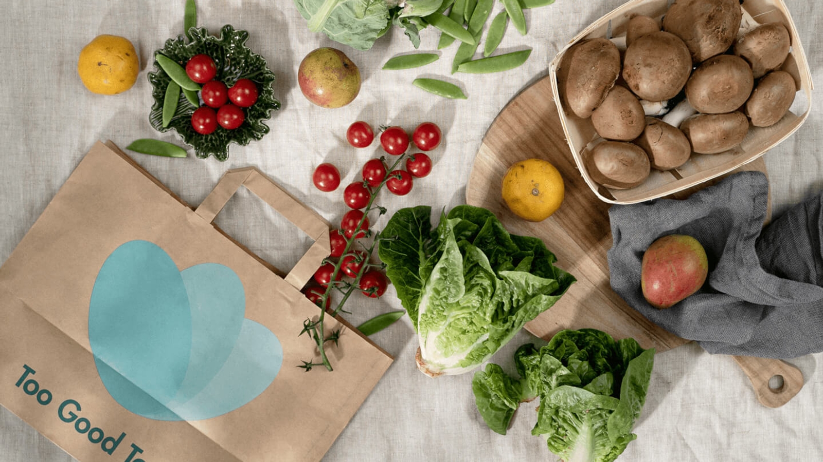 A collection of fresh vegetables including lettuce, mushrooms, and tomatoes next to a brown paper bag with the 'Too Good To Go' logo, indicating food rescue efforts.