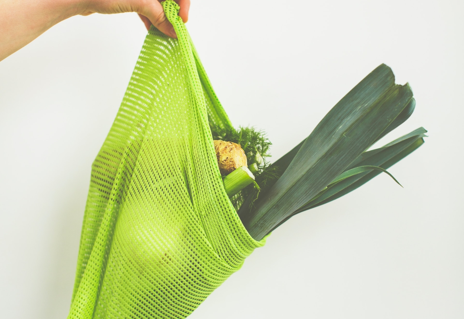A hand holding a bright green reusable mesh grocery bag containing leeks and a celery root, set against a white background.