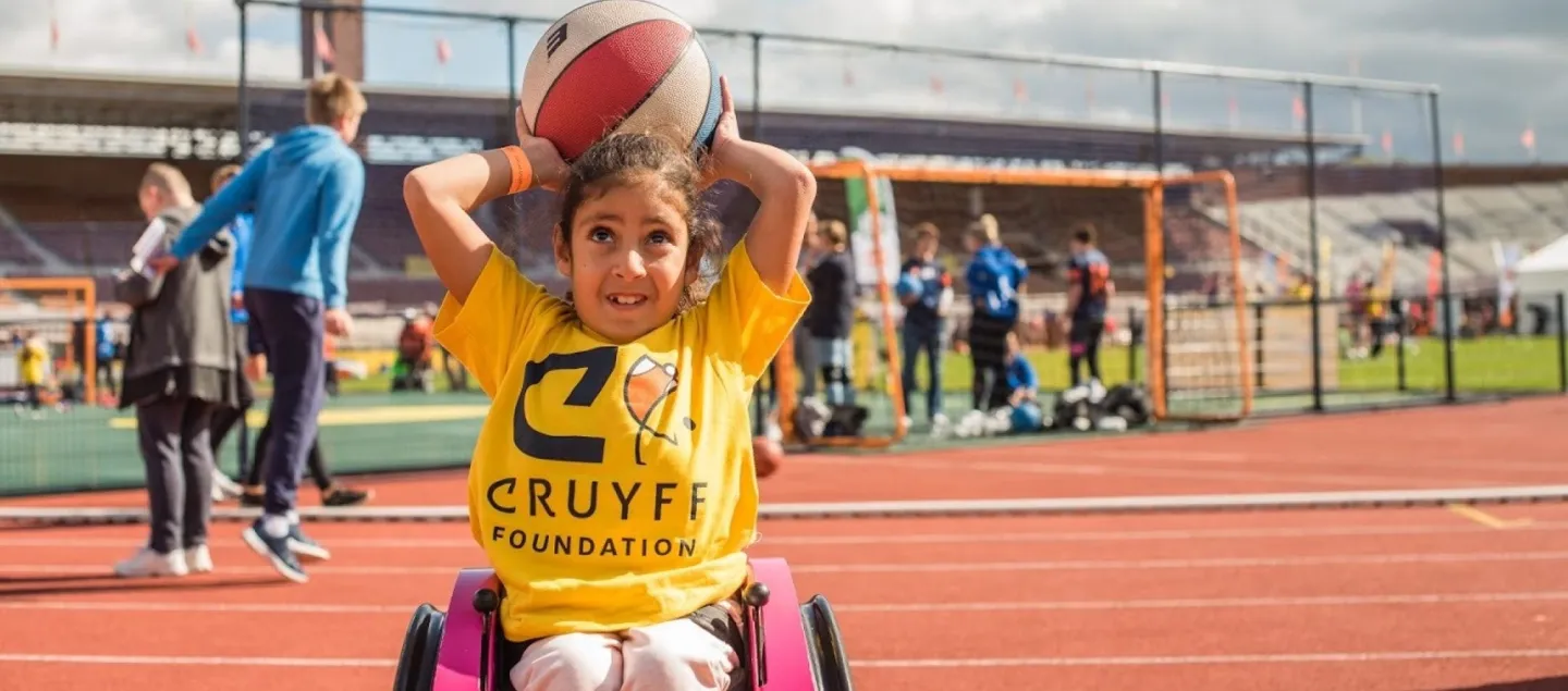 Meisje in Cruyff Foundation t-shirt speelt basketbal vanuit een rolstoel op een Cruyff Court