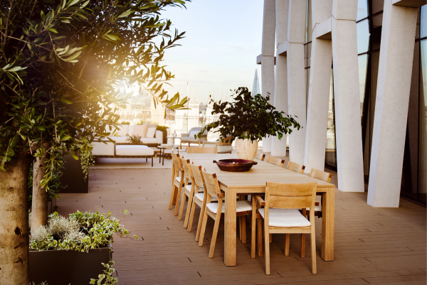 Expansive roof terrace with wooden dining table, cushioned chairs, sculptural planting and panoramic views over London, including The Shard and Westminster skyline.