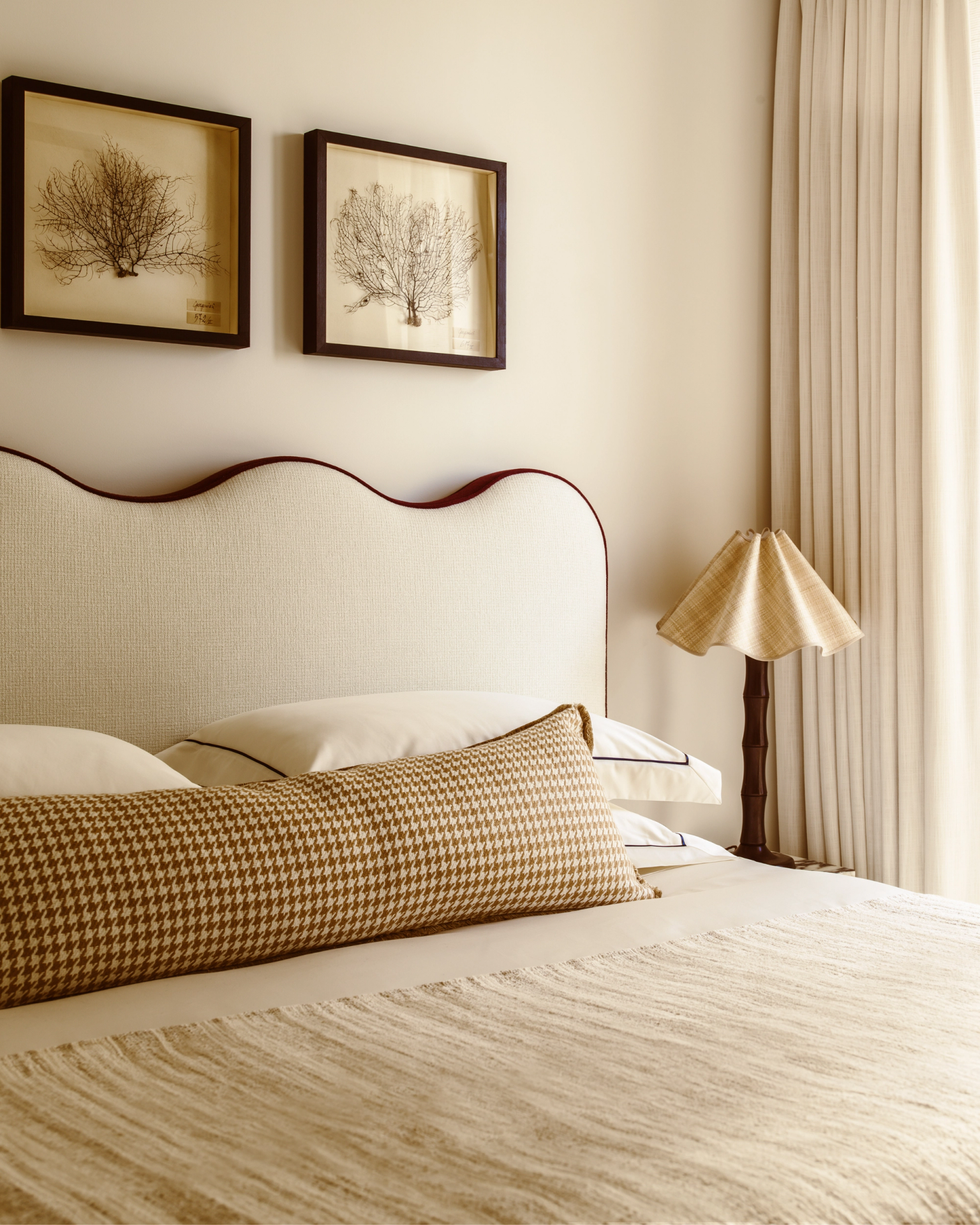 Serene bedroom with cream scalloped headboard, houndstooth bolster cushion, pleated lamp and botanical artwork, designed in a warm neutral palette.