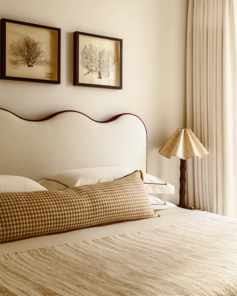 Serene bedroom with cream scalloped headboard, houndstooth bolster cushion, pleated lamp and botanical artwork, designed in a warm neutral palette.