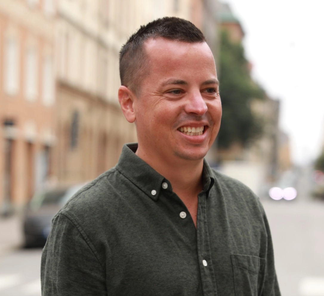 Den leendes coachen och föreläsaren Simon Beyer i mörkgrön skjorta med hus och gata som bakgrund